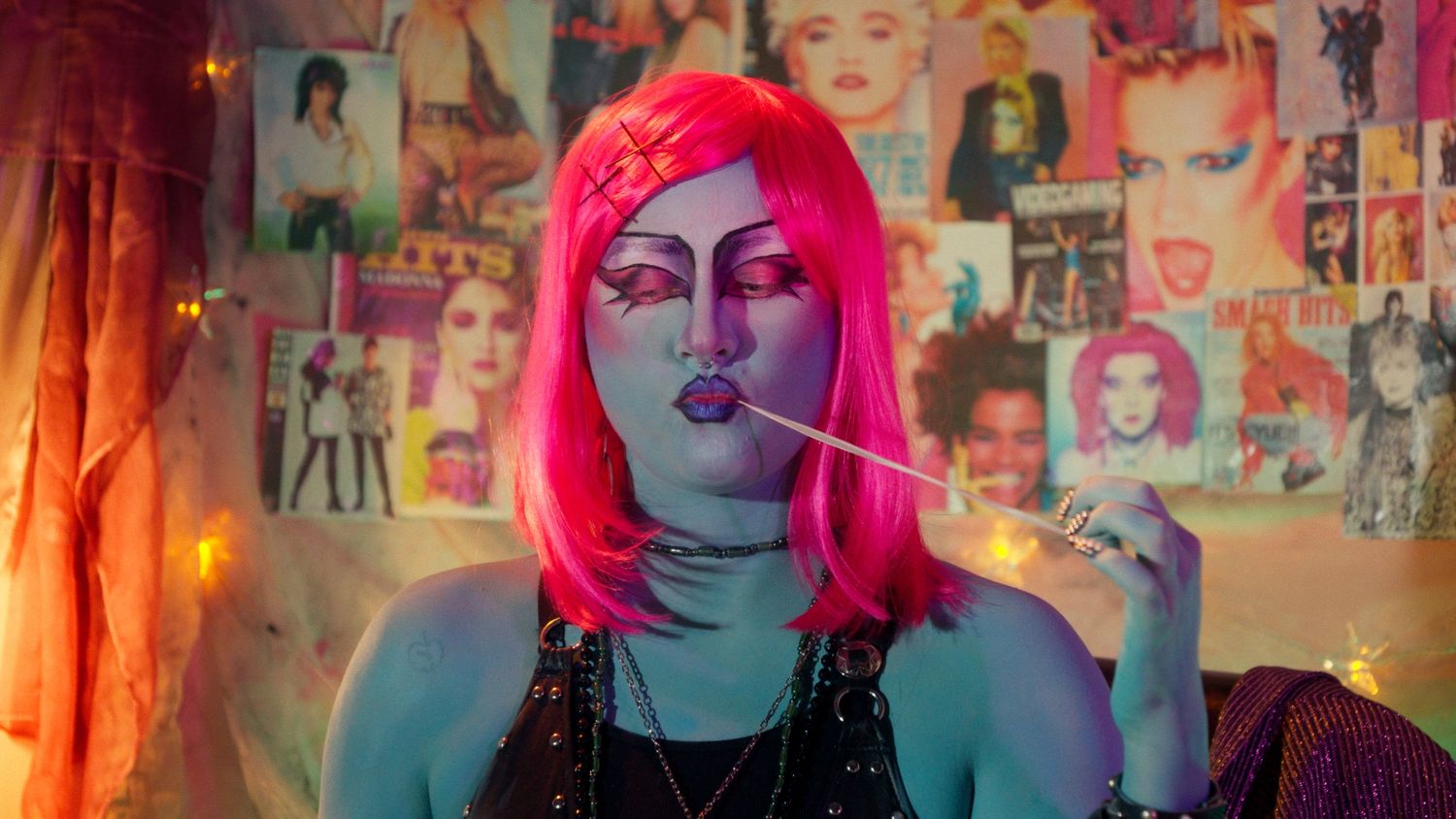 Close-up of a person with pink wig and blue face makeup holding a lollipop in front of pop culture posters.