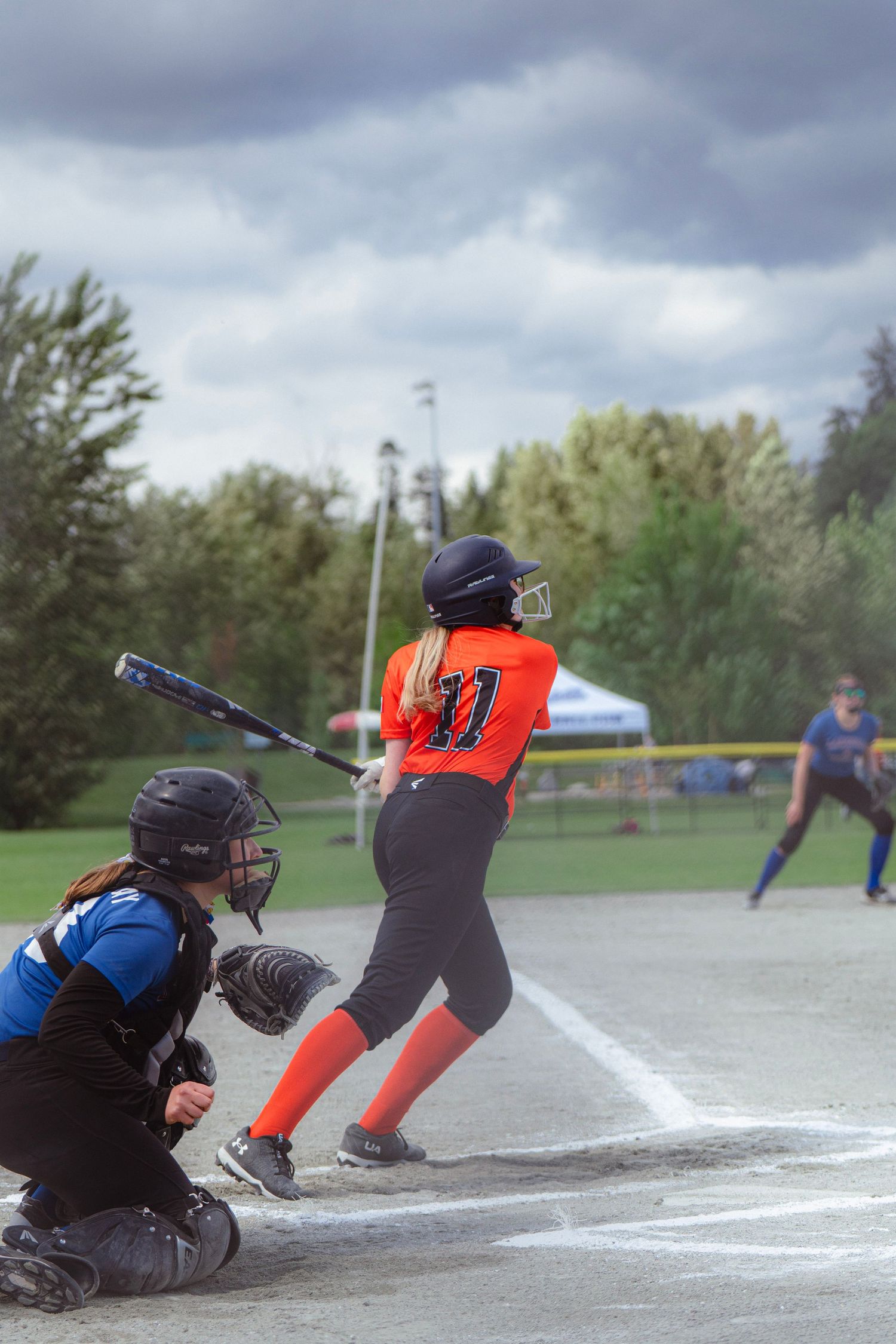 Softball batter in orange uniform swings at pitch as catcher crouches behind home plate on cloudy day.
