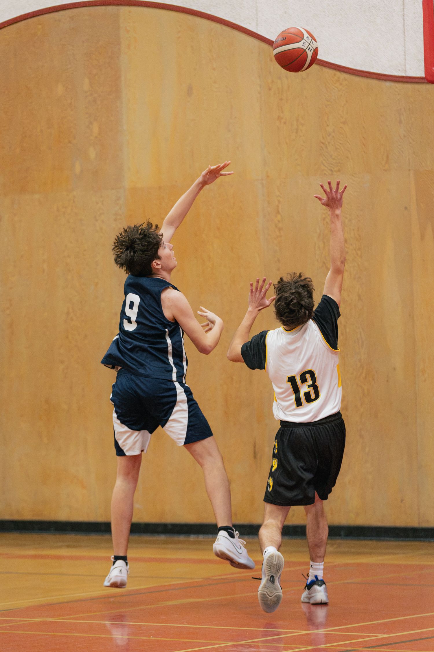 Player #9 in navy jersey releases a jump shot over defender #13 in white during an indoor basketball game.
