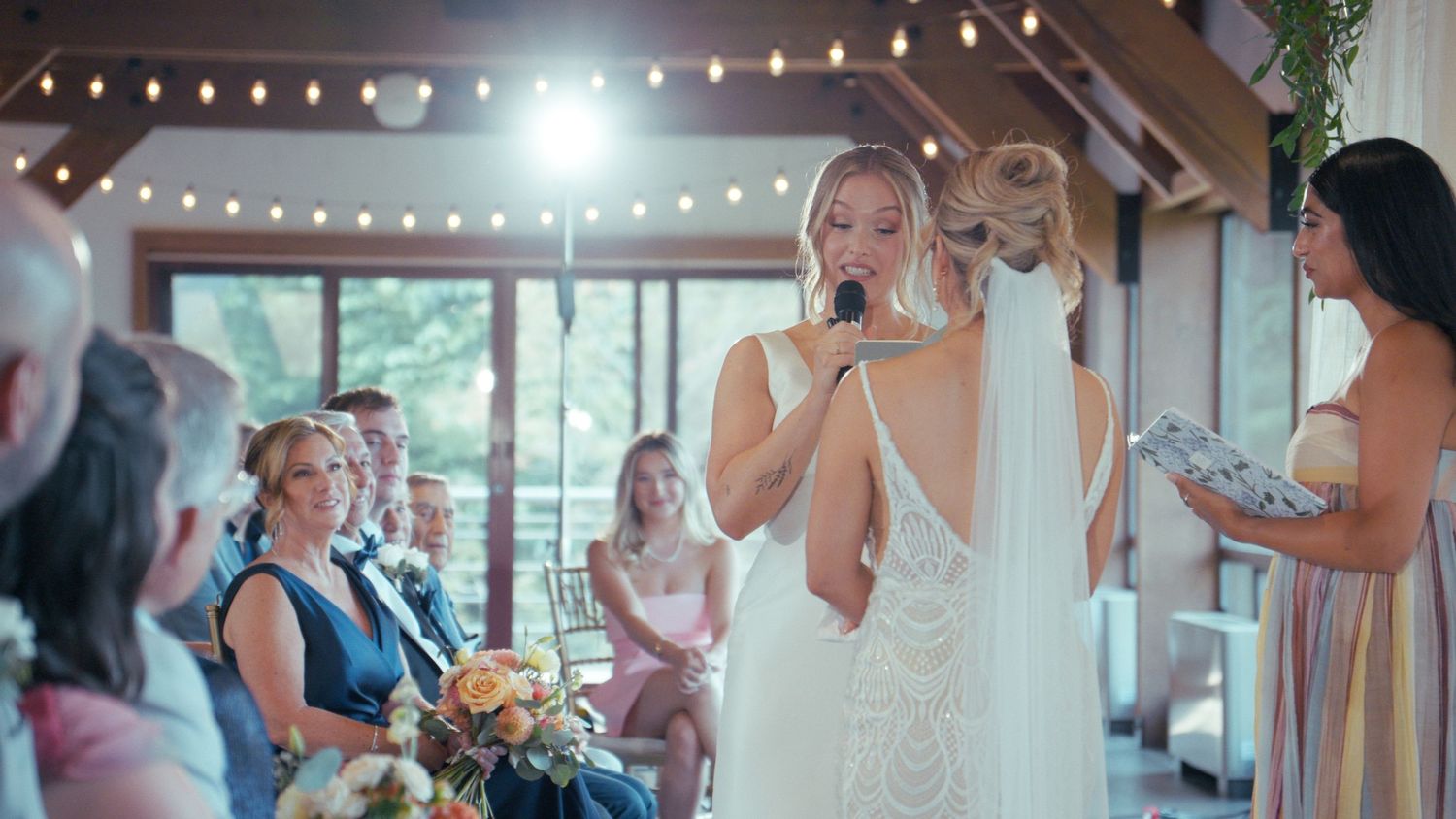 Blonde bride in backless lace gown reads vows into microphone at rustic barn wedding ceremony.