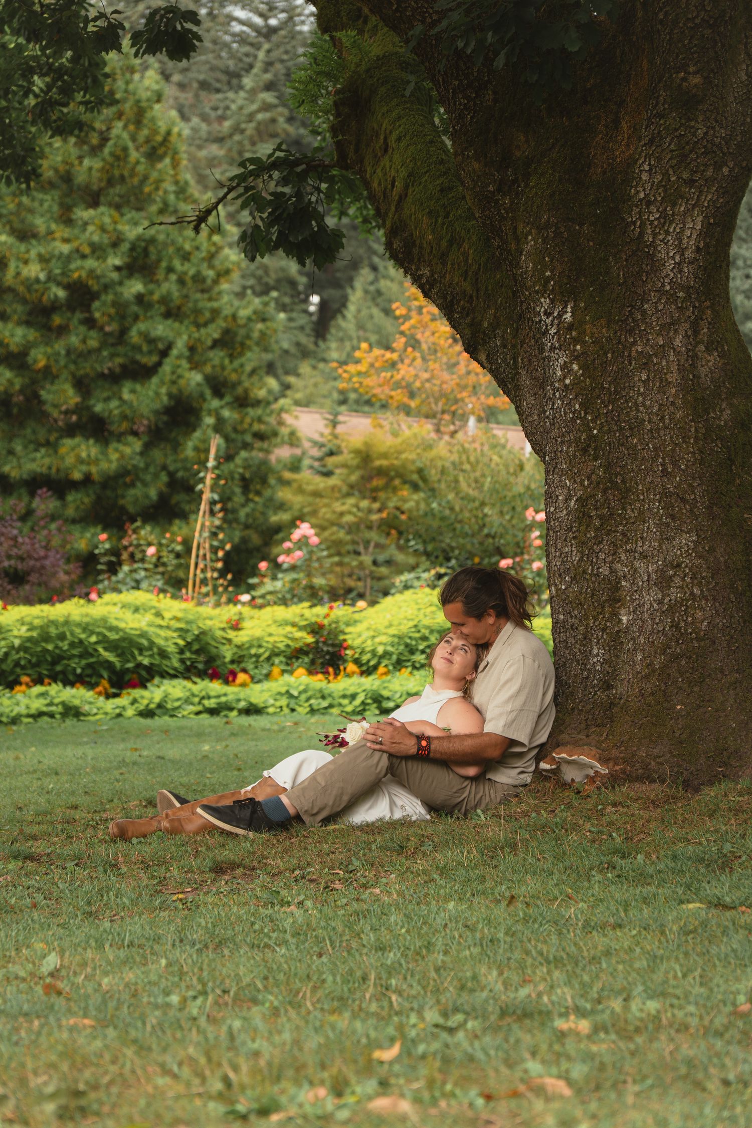 Couple sitting together under a large tree in a lush green garden, sharing a romantic embrace.