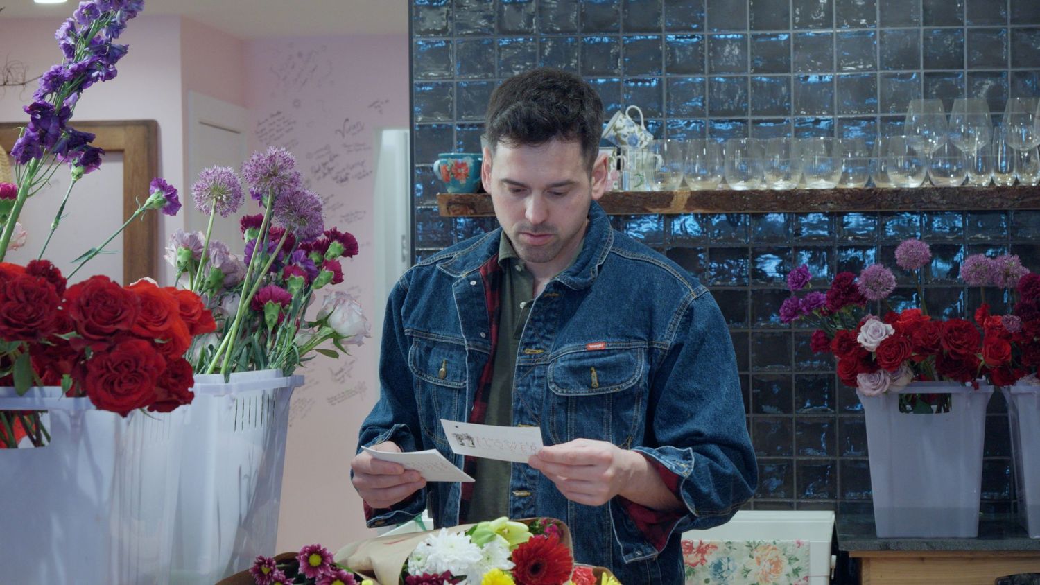 Young man in denim jacket reads cards while standing among colorful flower arrangements in a floral shop.