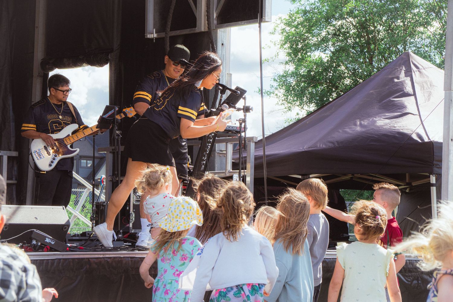 Live band performer leans toward crowd of young children gathered at outdoor festival stage.