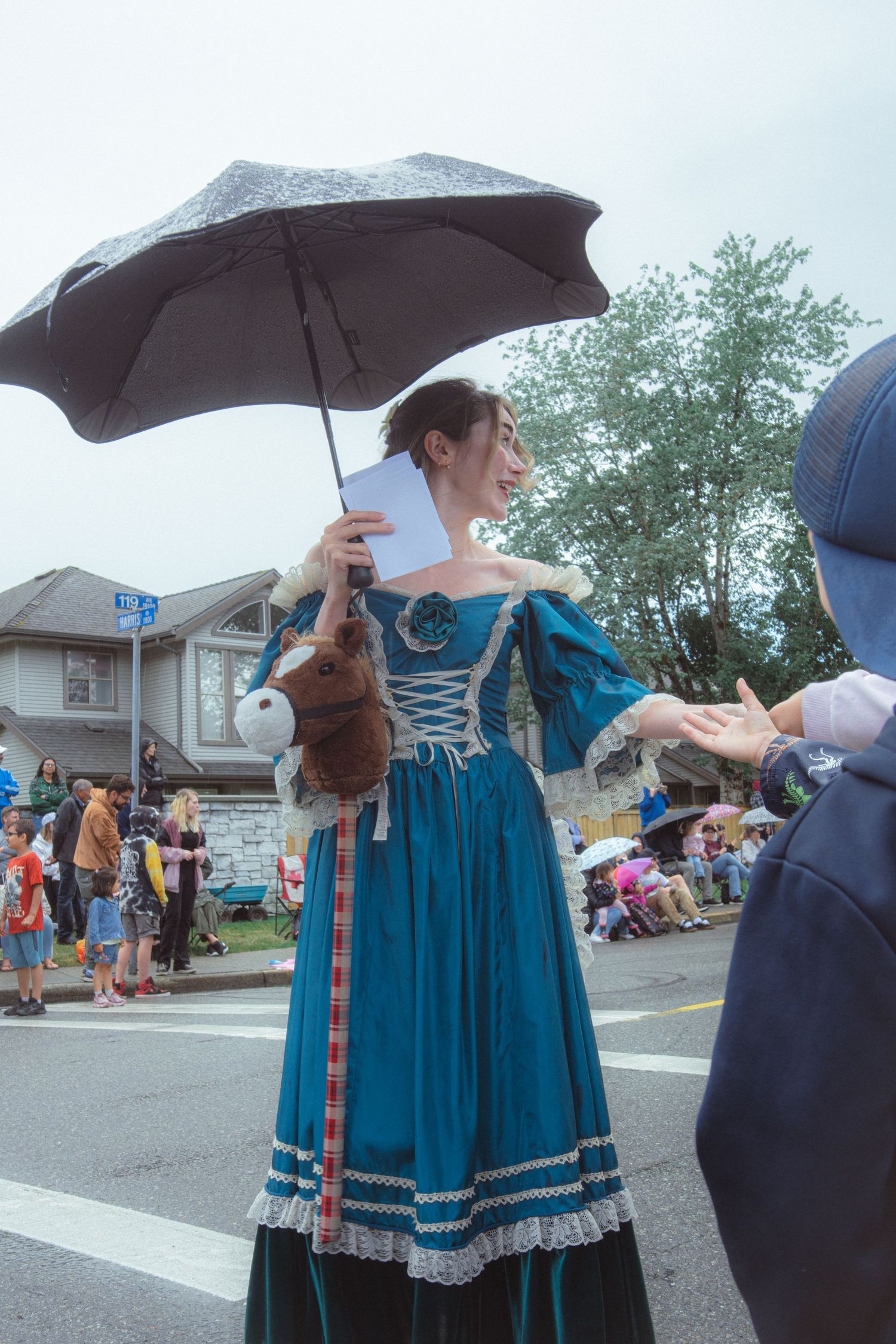 Woman in teal Victorian dress holding umbrella and stuffed animal at rainy outdoor parade.