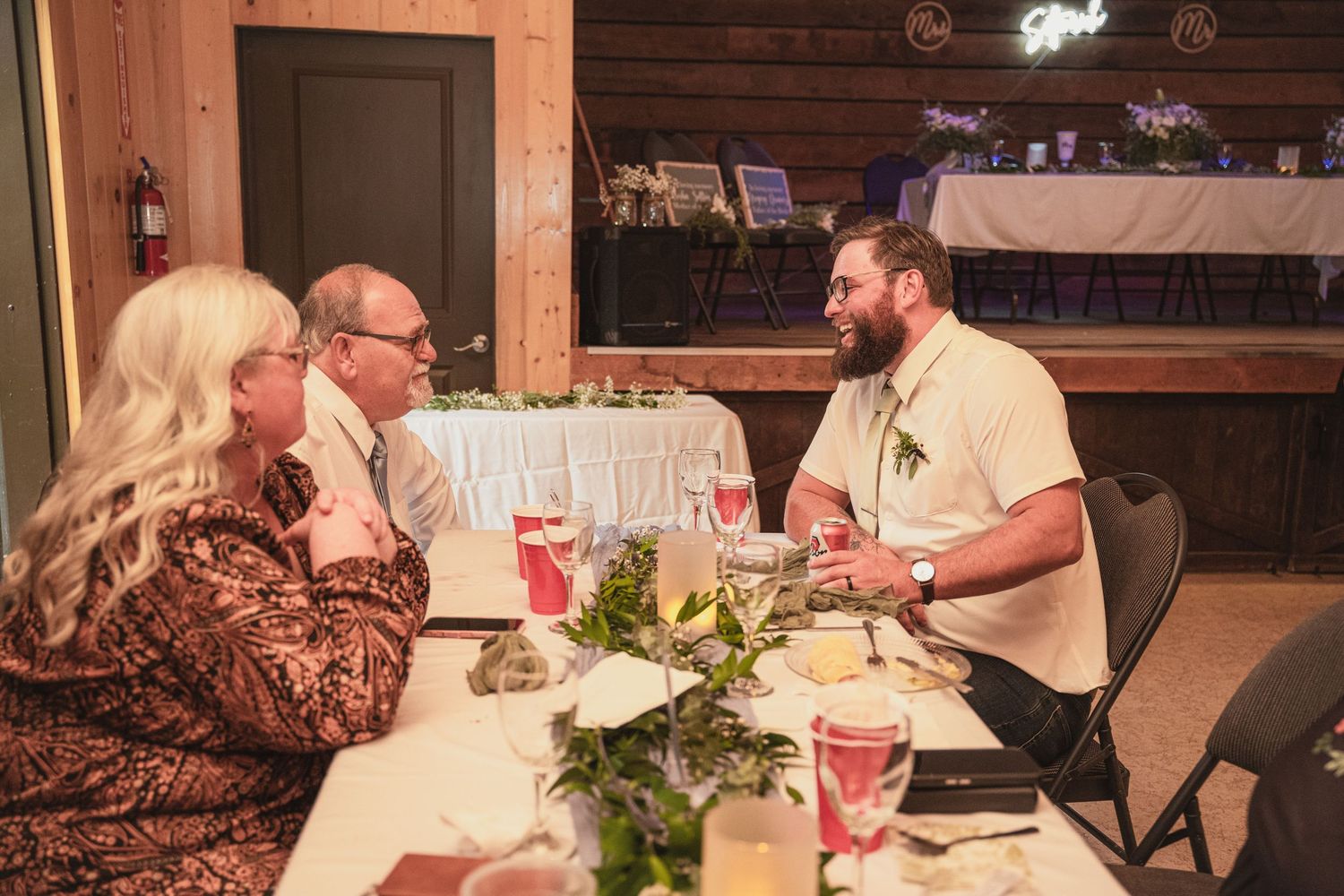 Bearded groom in white shirt chats with seated wedding guests at an elegantly set reception table.