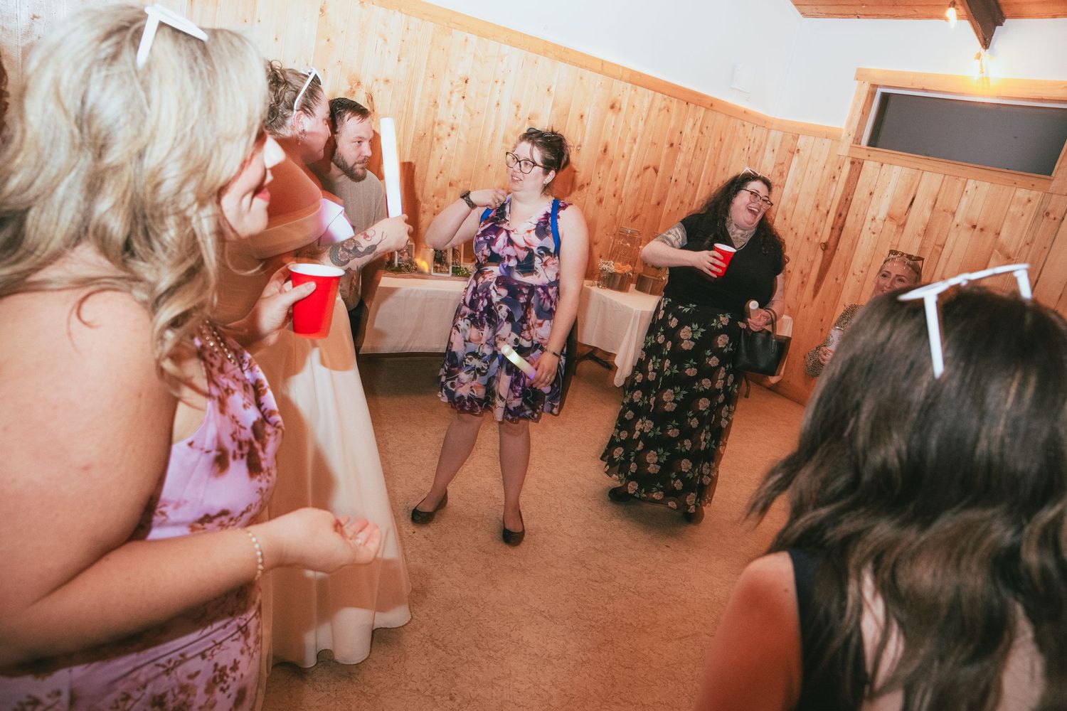 Women dancing and celebrating at a rustic indoor wedding reception with wood-paneled walls.