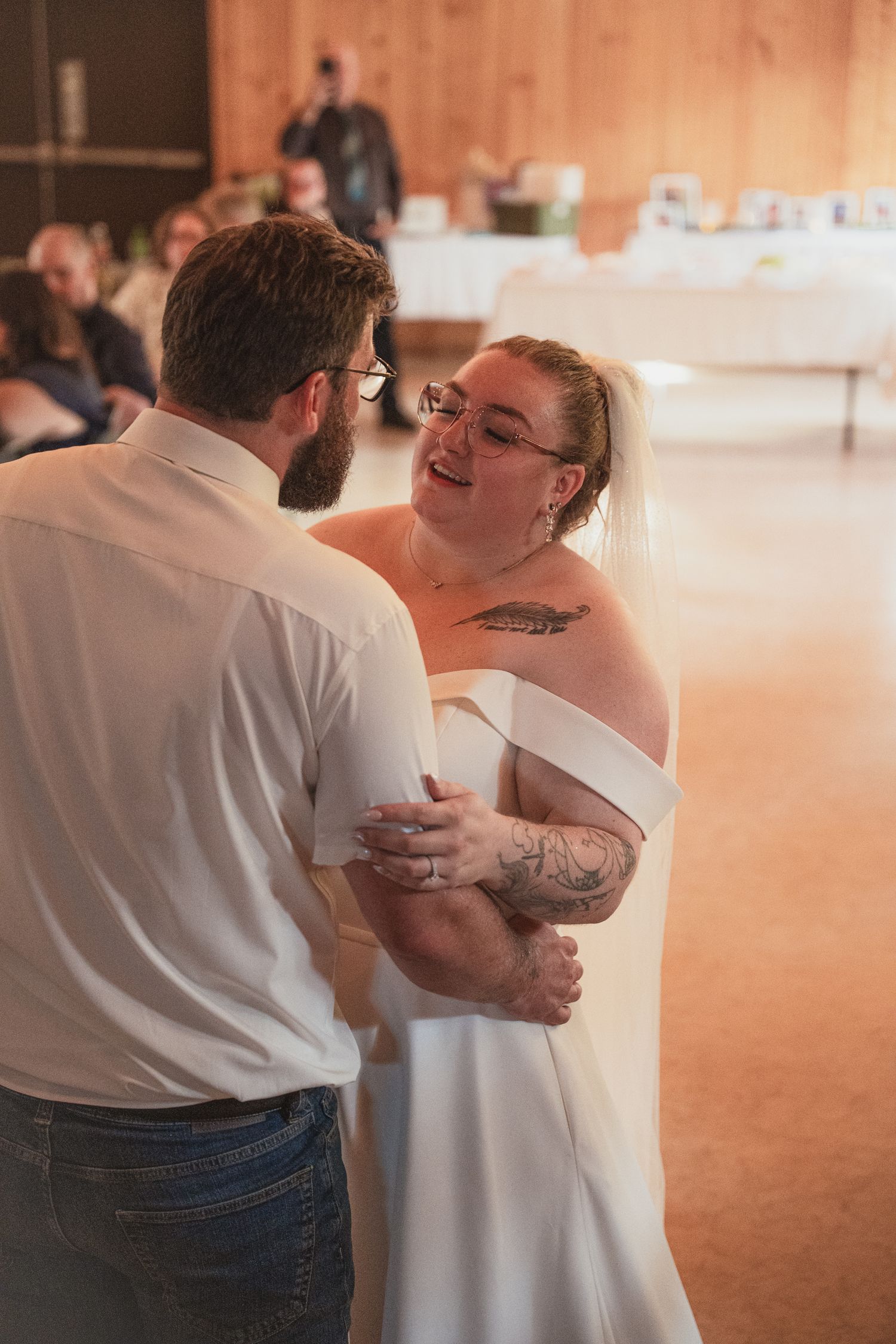 Bride in white off-shoulder gown with tattoos shares first dance with groom in white shirt at rustic wedding reception.