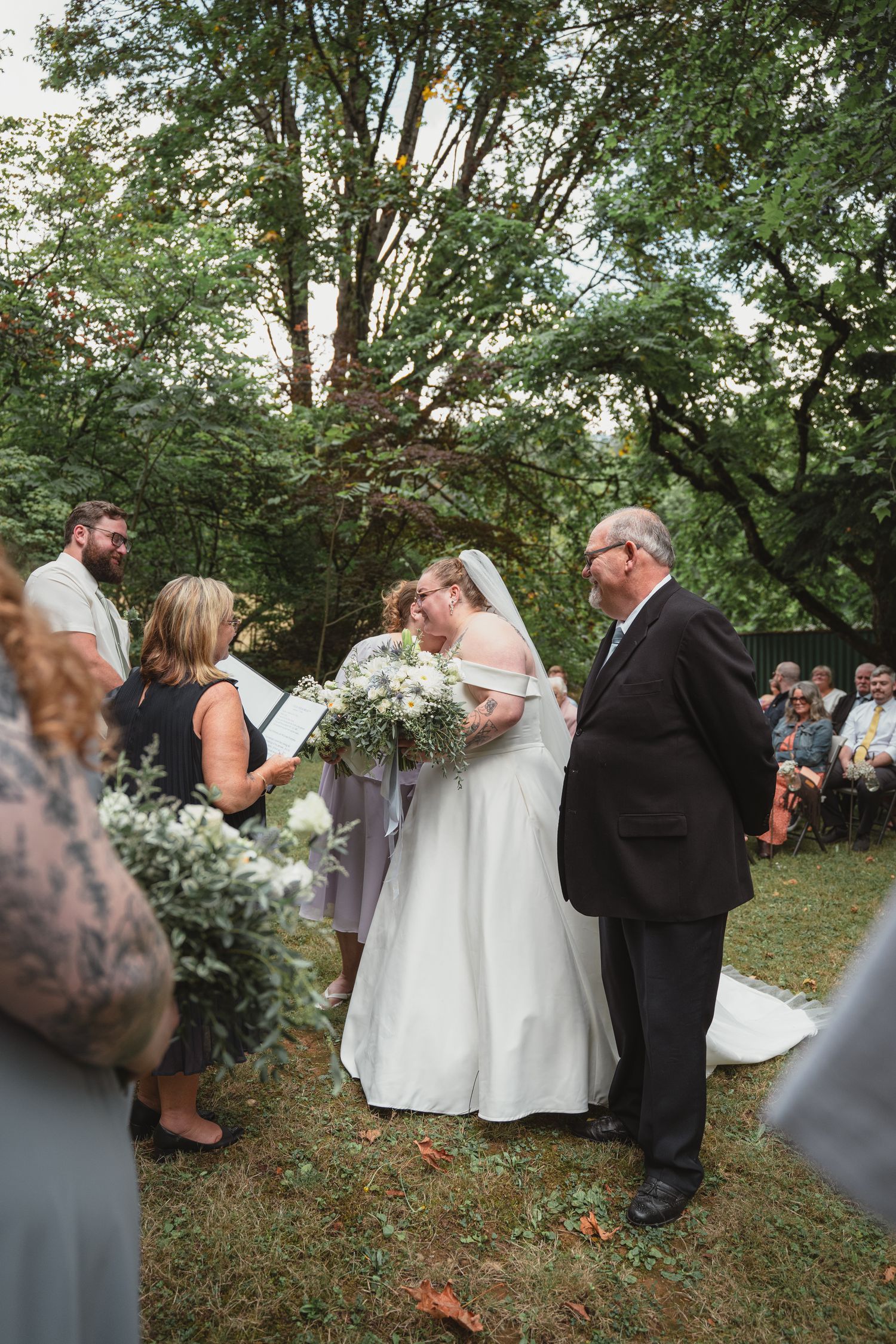 Outdoor wedding ceremony with bride in white gown holding bouquet, officiant reading vows, surrounded by lush green trees.
