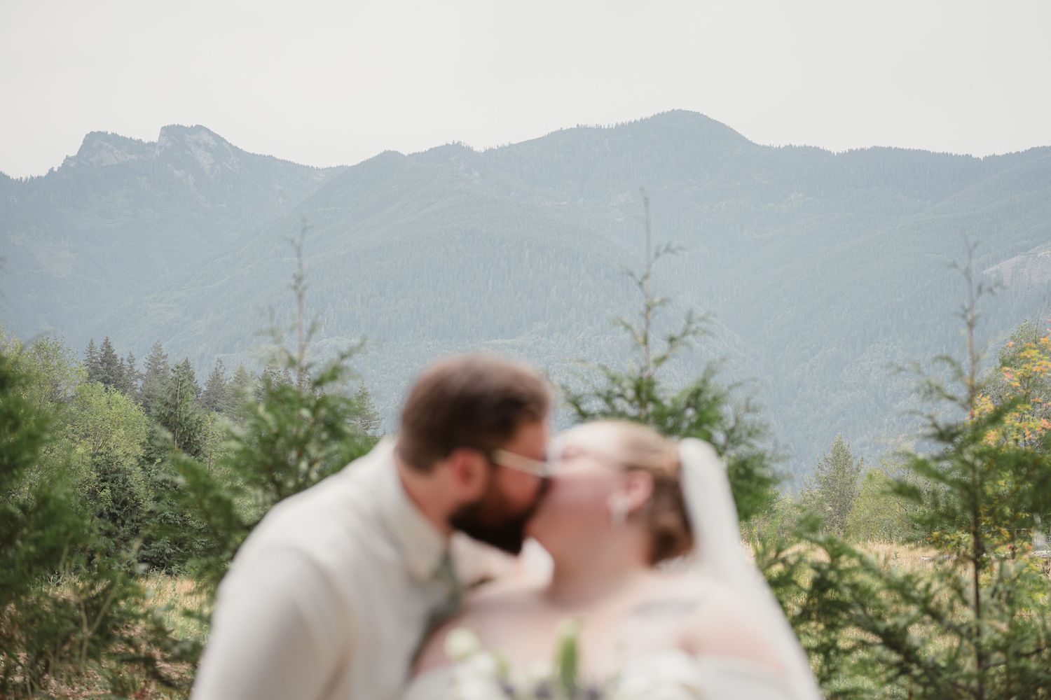 A couple shares a kiss with misty mountain peaks and evergreen trees in the background at their outdoor wedding.