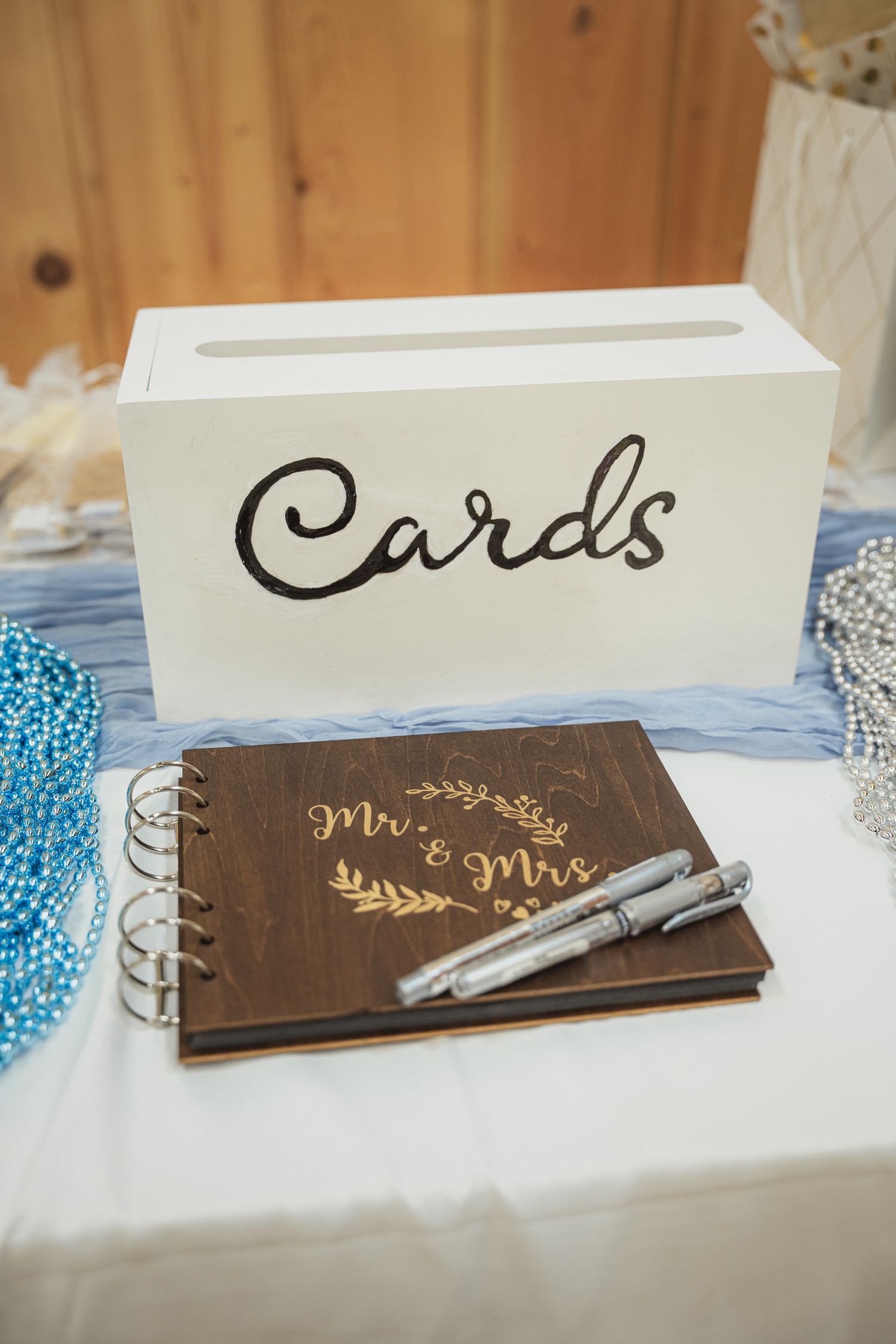 Rustic wedding guest book engraved with Mr. & Mrs. beside a white Cards box with pens on a decorated table.
