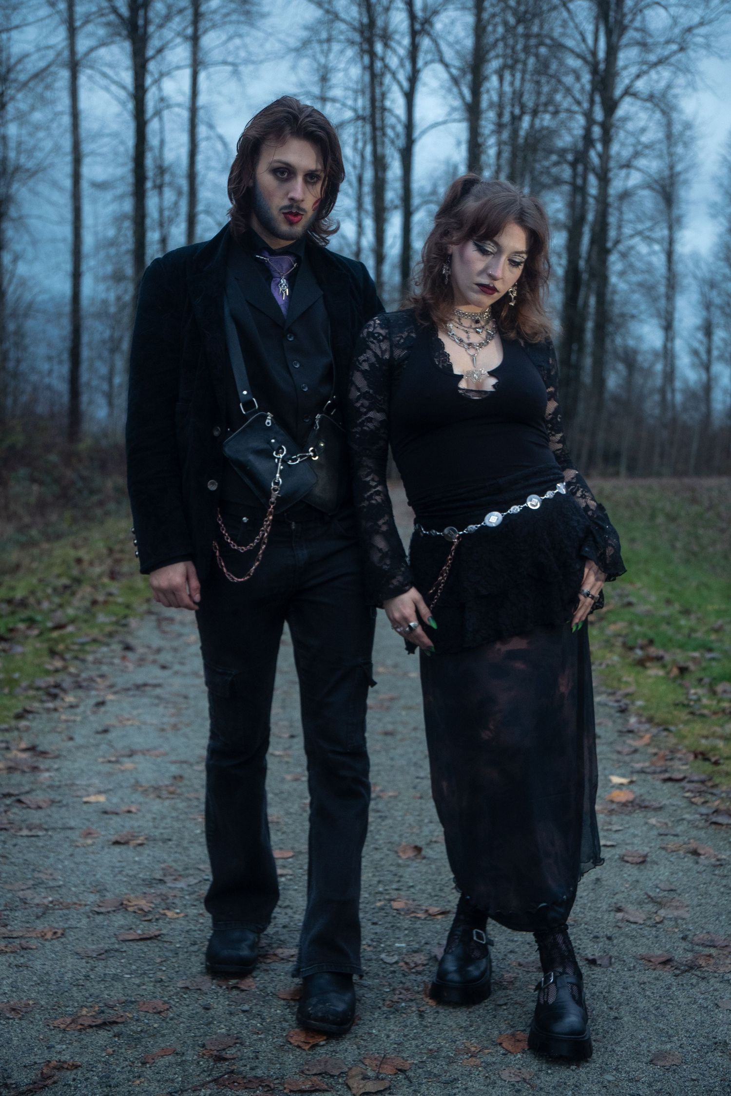 A gothic couple dressed in all-black Victorian-inspired outfits walk together on a forest path at dusk.