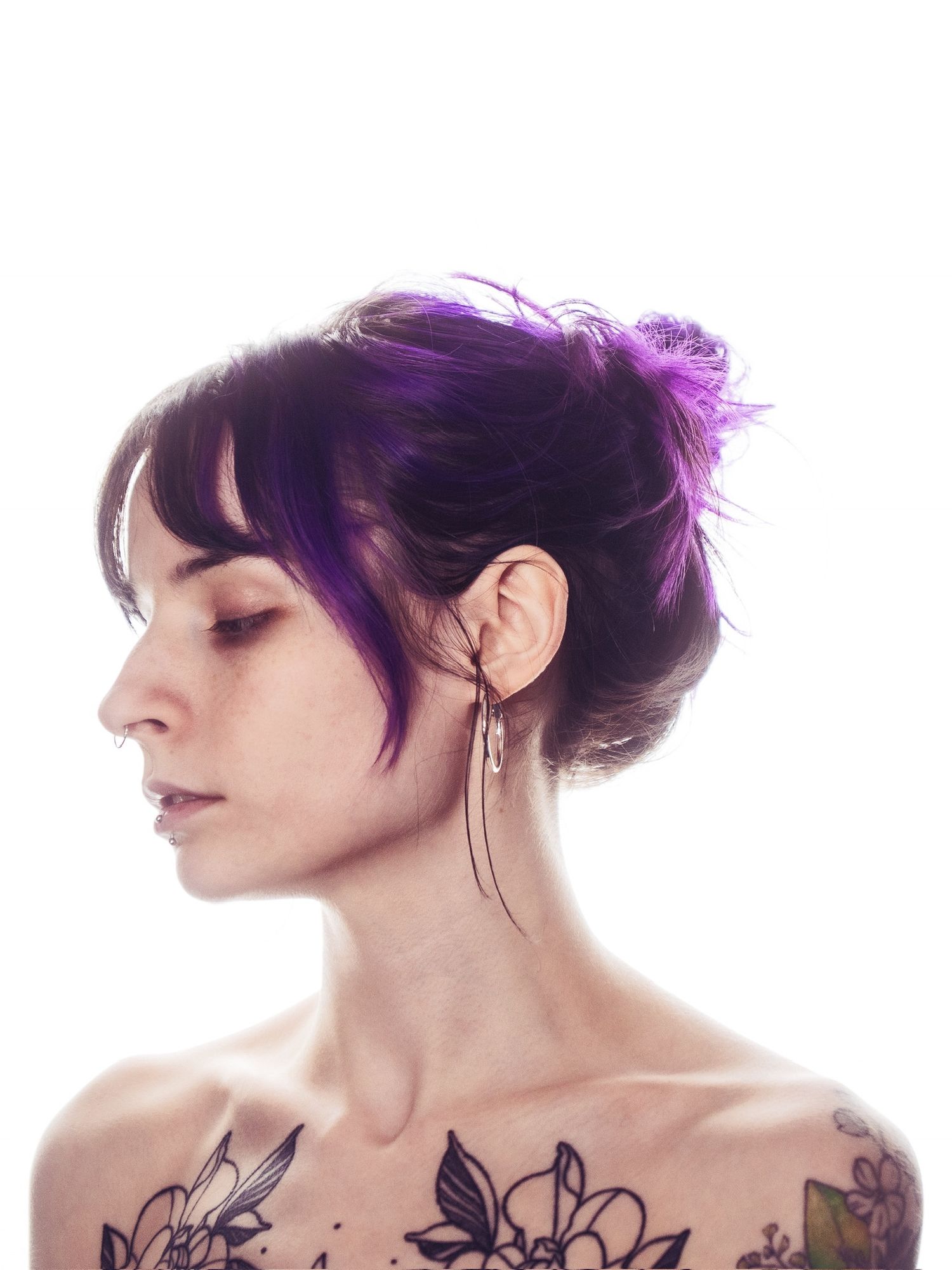 Side profile of woman with purple updo hair and floral tattoos on shoulders wearing dangling earring.