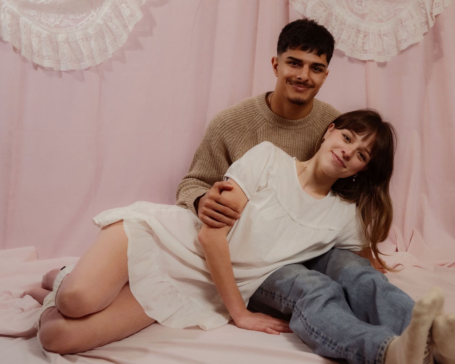 Young couple sitting together on a pink surface, man in beige sweater embracing woman in white dress.