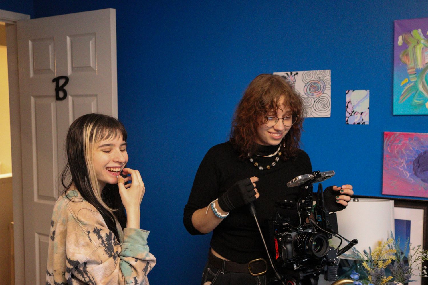 Filmmaker and actress share a laugh on set with blue wall and artwork in background.