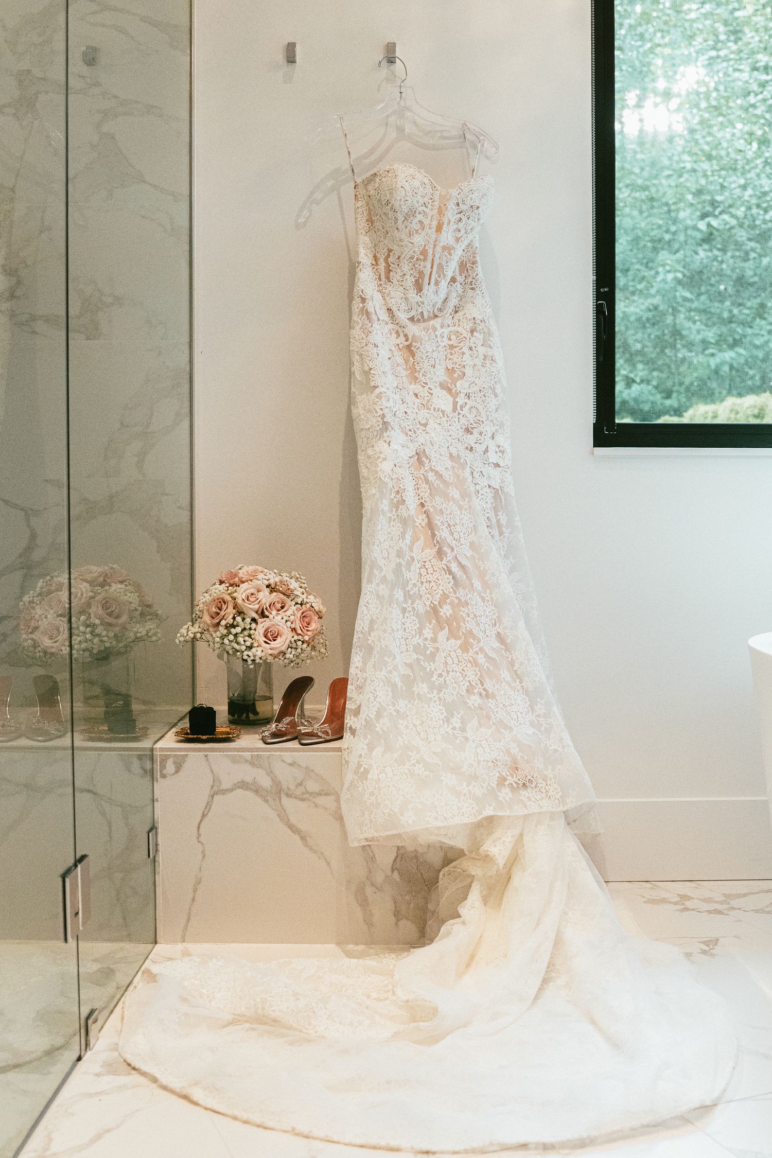 Stunning lace wedding dress with long train hanging in a marble bathroom beside pink bridal bouquet and heels.