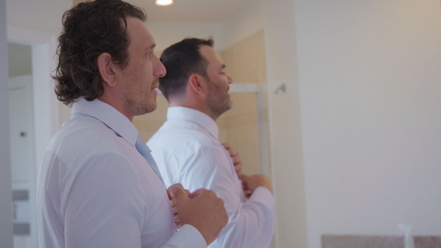 Two men in white dress shirts adjusting their collars in a bright room, preparing for a formal wedding event.