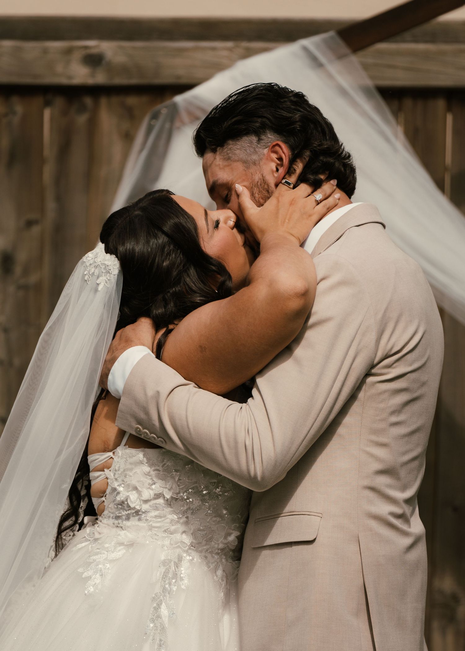 Bride and groom share their first kiss at the altar, bride in lace gown with veil, groom in beige suit.