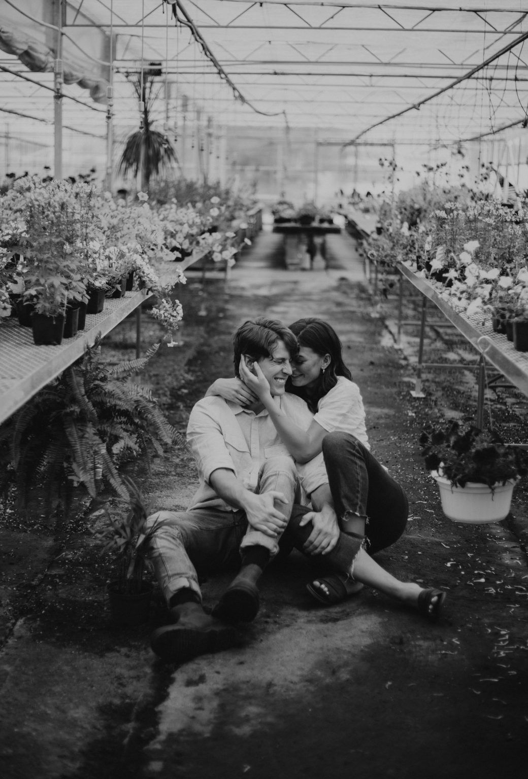 A couple sits intimately on the ground in a greenhouse surrounded by blooming plants in a black and white photo.