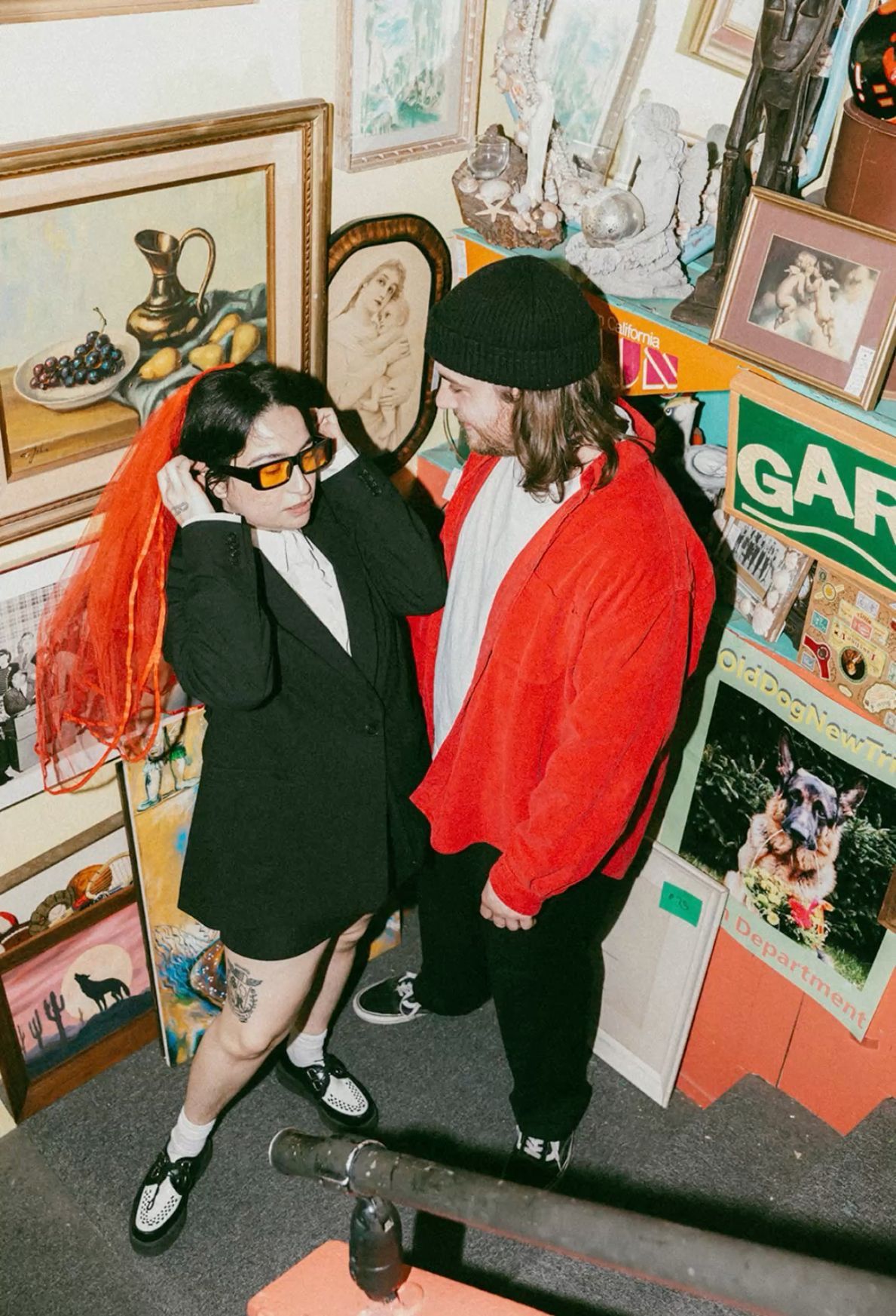 Two stylish people pose in an art-filled shop, one wearing orange sunglasses and red hair, the other in a red jacket.