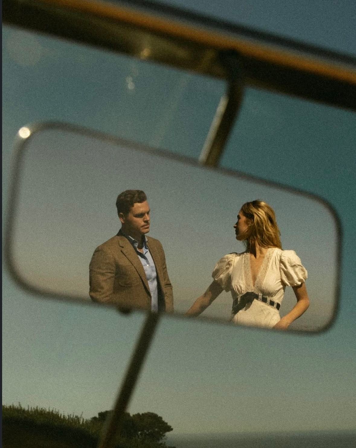 A man and woman reflected in a vintage car rearview mirror against a twilight sky.