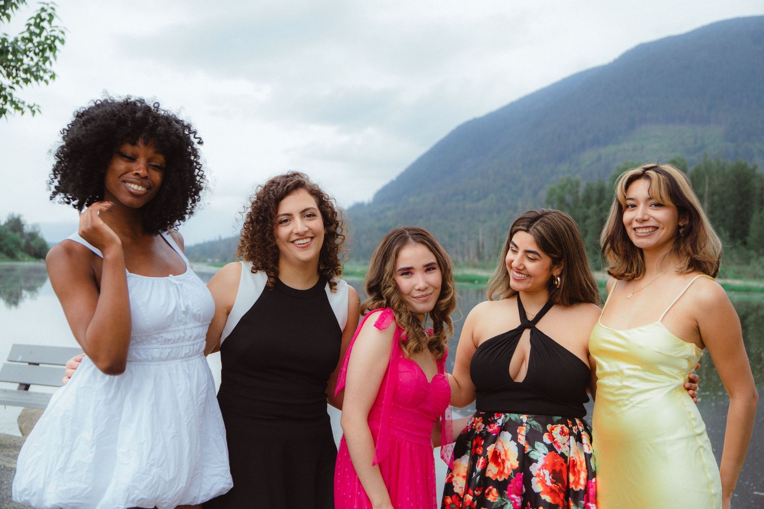 Five smiling women in elegant dresses pose together outdoors with a mountain lake in the background.