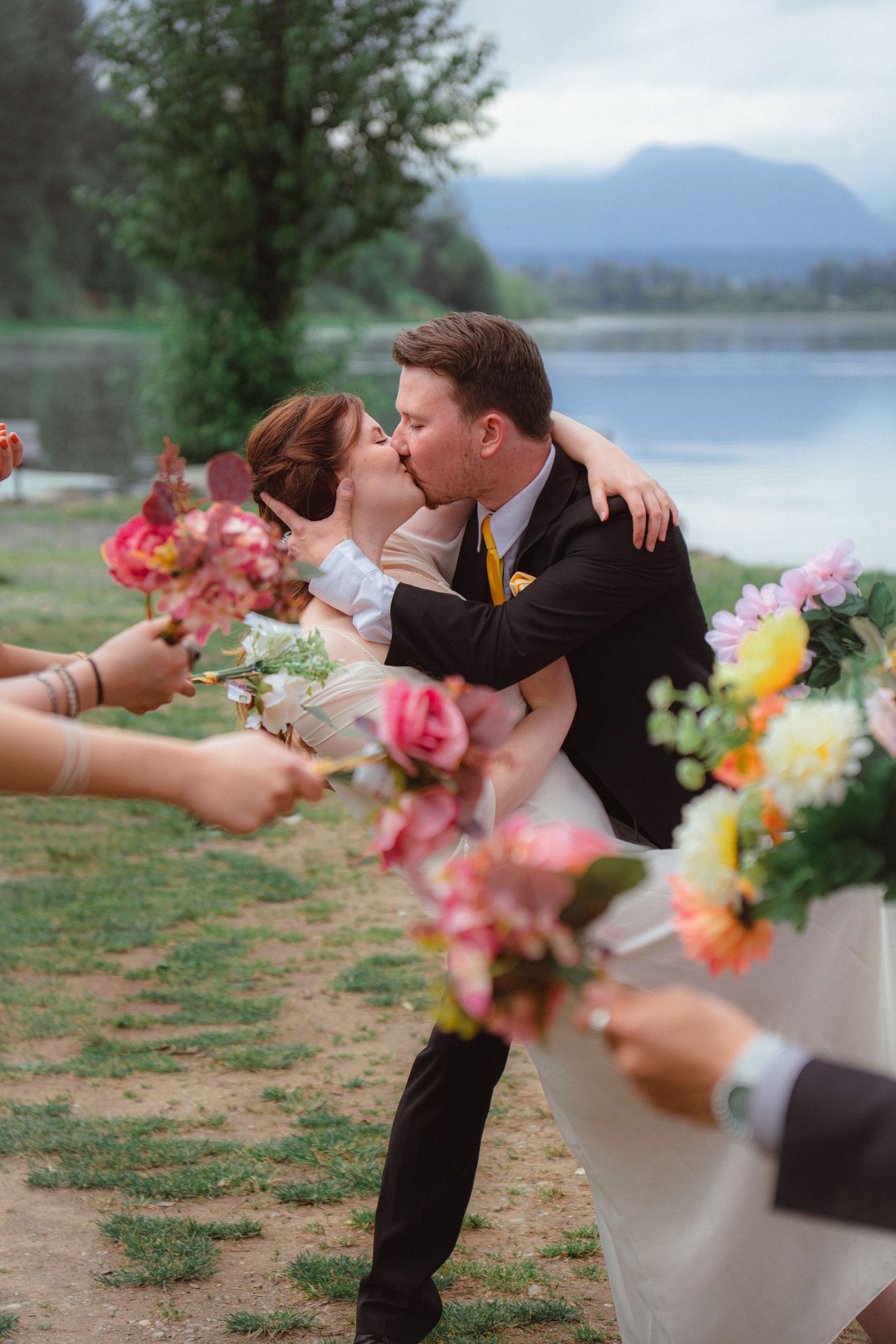 Newlyweds sharing romantic kiss surrounded by guests holding flowers at lakeside wedding ceremony.