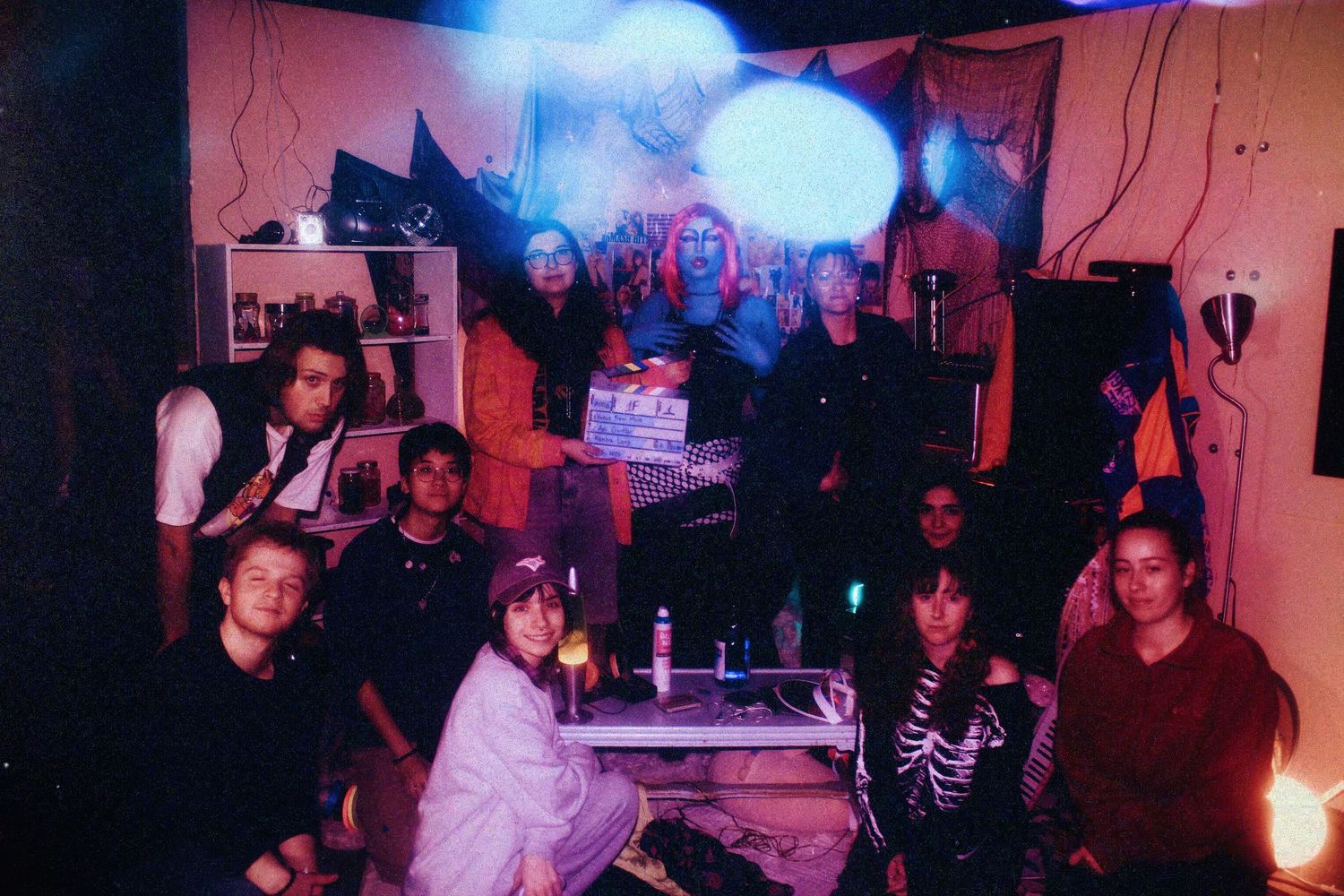 Group of film students pose together on a vibrant neon-lit movie set holding a clapperboard.