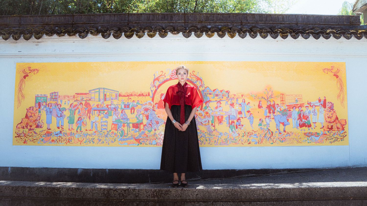 Woman in black dress stands before vibrant yellow mural depicting colorful crowd scene outdoors.