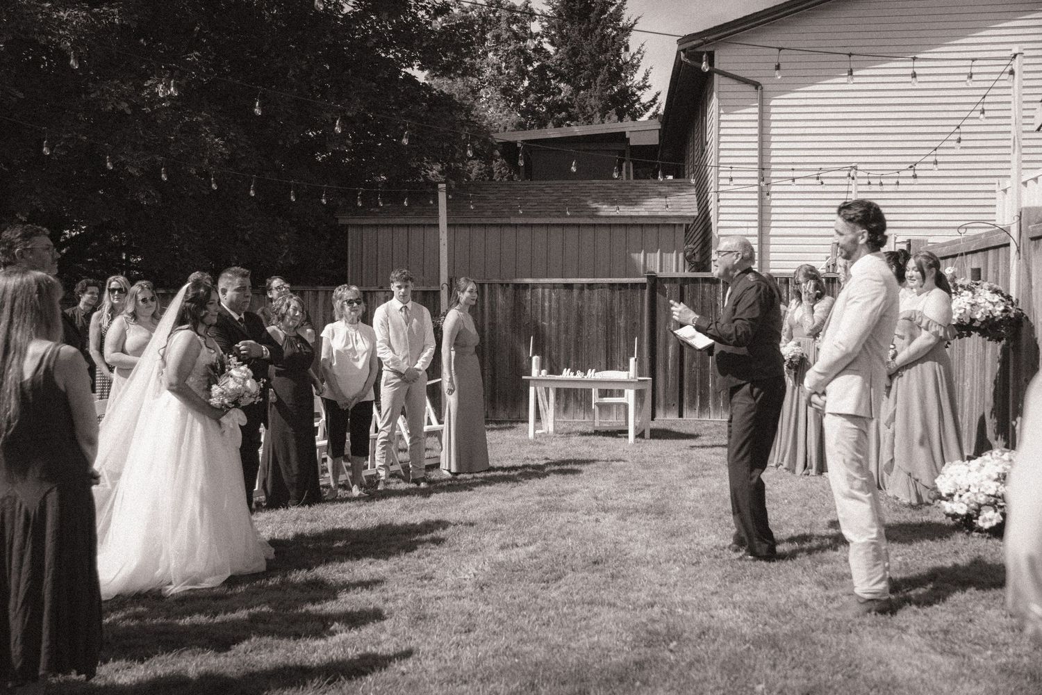 Black and white outdoor backyard wedding ceremony with bride, groom, and officiant on sunny day.