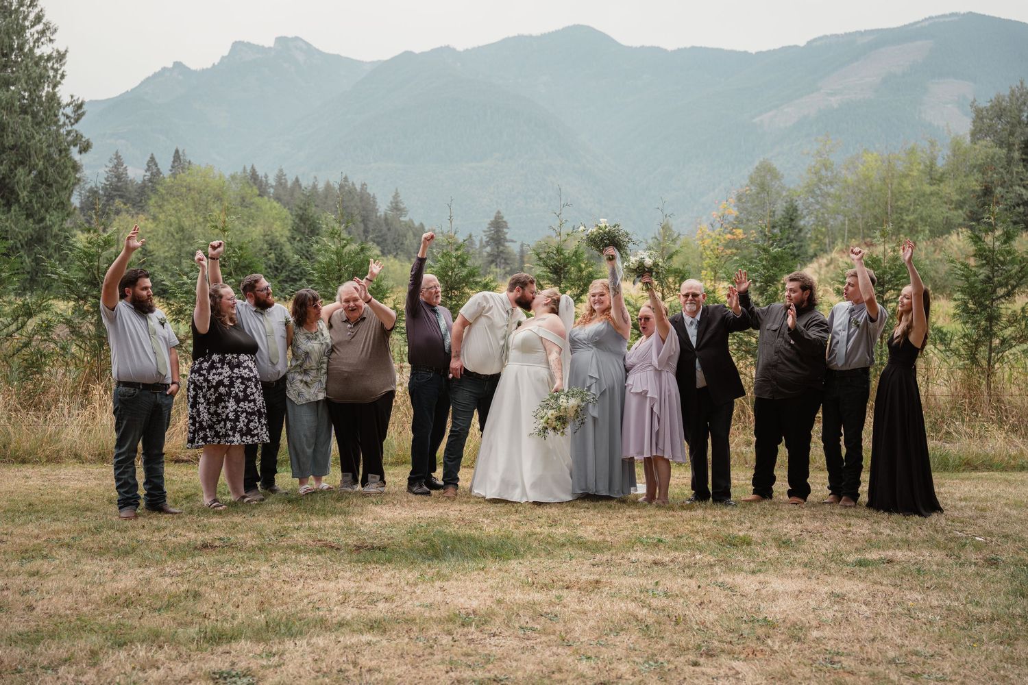 Wedding party celebrating outdoors with mountains and evergreen trees in the background, bride and groom surrounded by guests.