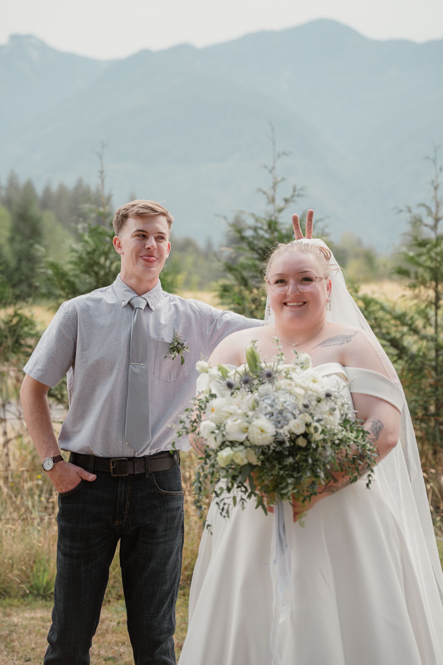 Bride holding white bouquet stands with groomsman against mountain backdrop at outdoor wedding.