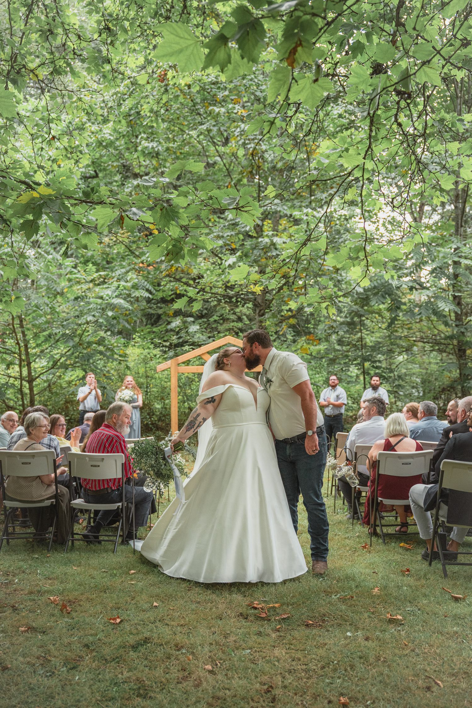Bride in white gown and groom share first kiss under wooden arch at lush outdoor wedding ceremony.