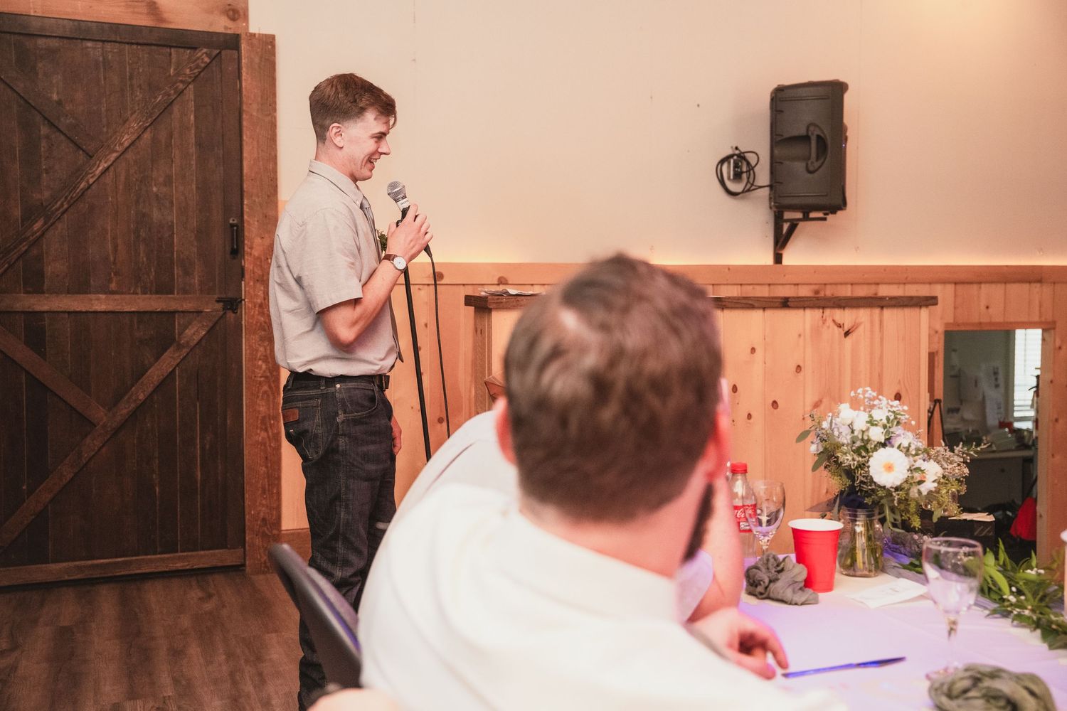 Young man delivers a heartfelt wedding speech into a microphone at a rustic barn reception venue.