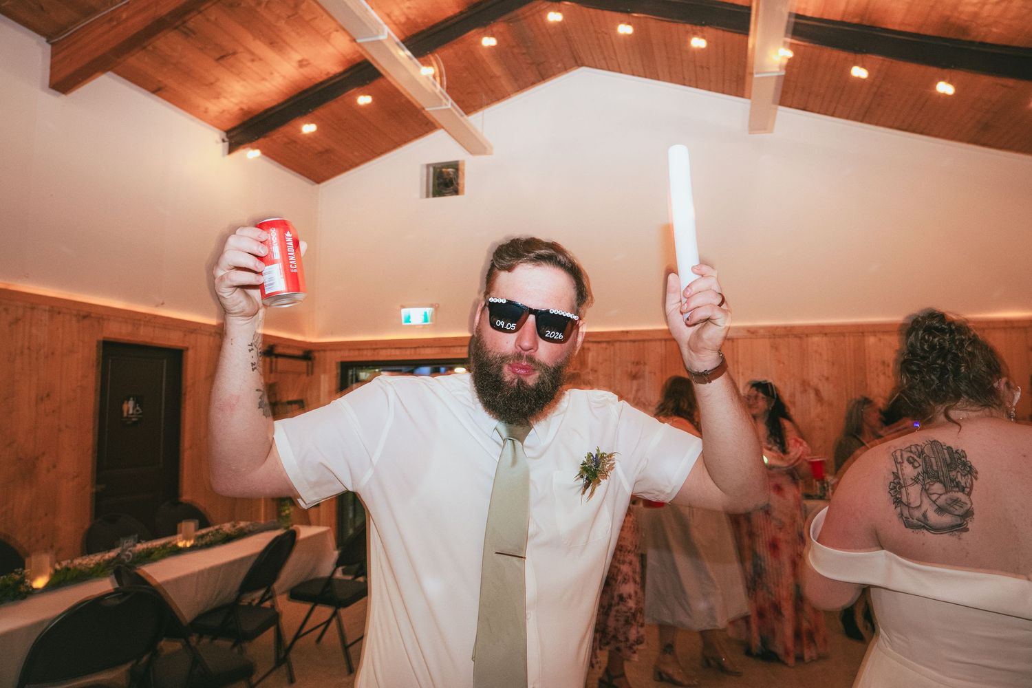 Bearded man in sunglasses celebrating at a wedding reception, holding a drink and glow stick in a wood-paneled venue.