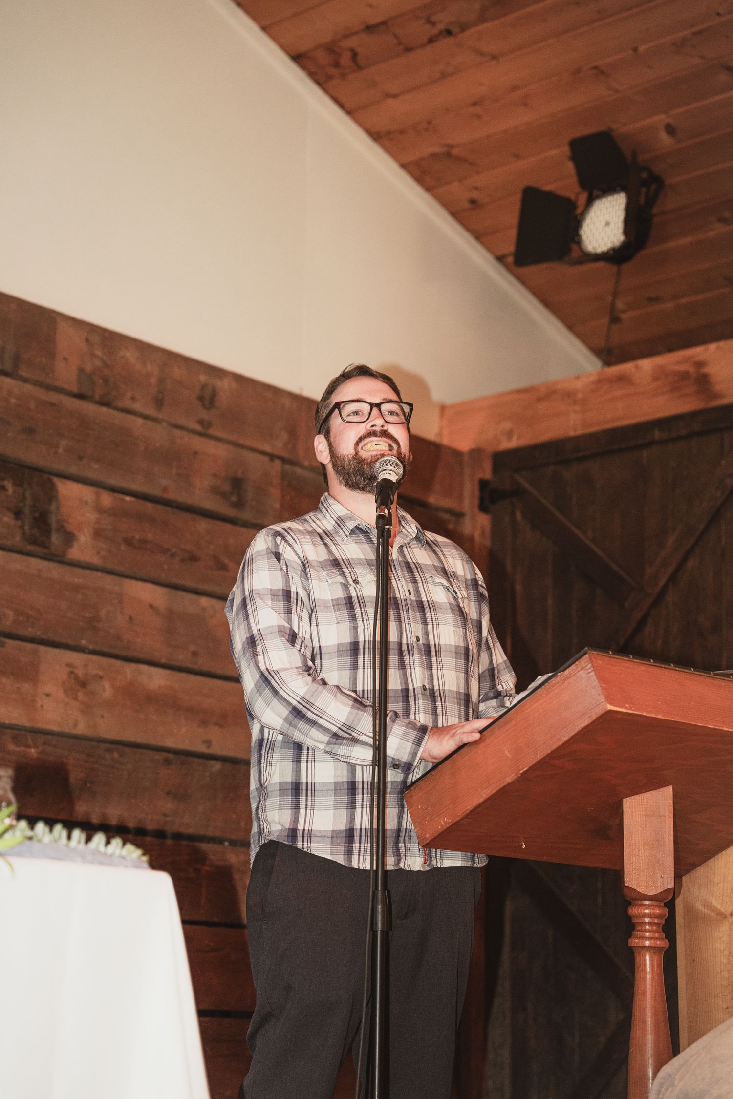 Man in plaid shirt speaking at wooden podium with microphone in rustic barn venue.