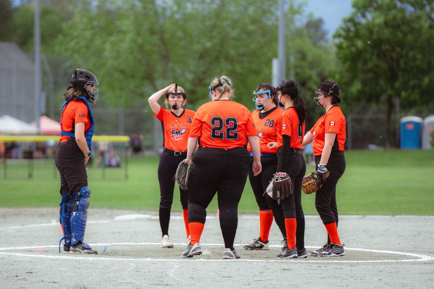 Female softball players in orange uniforms gather on the pitcher's mound for a team discussion during a game.