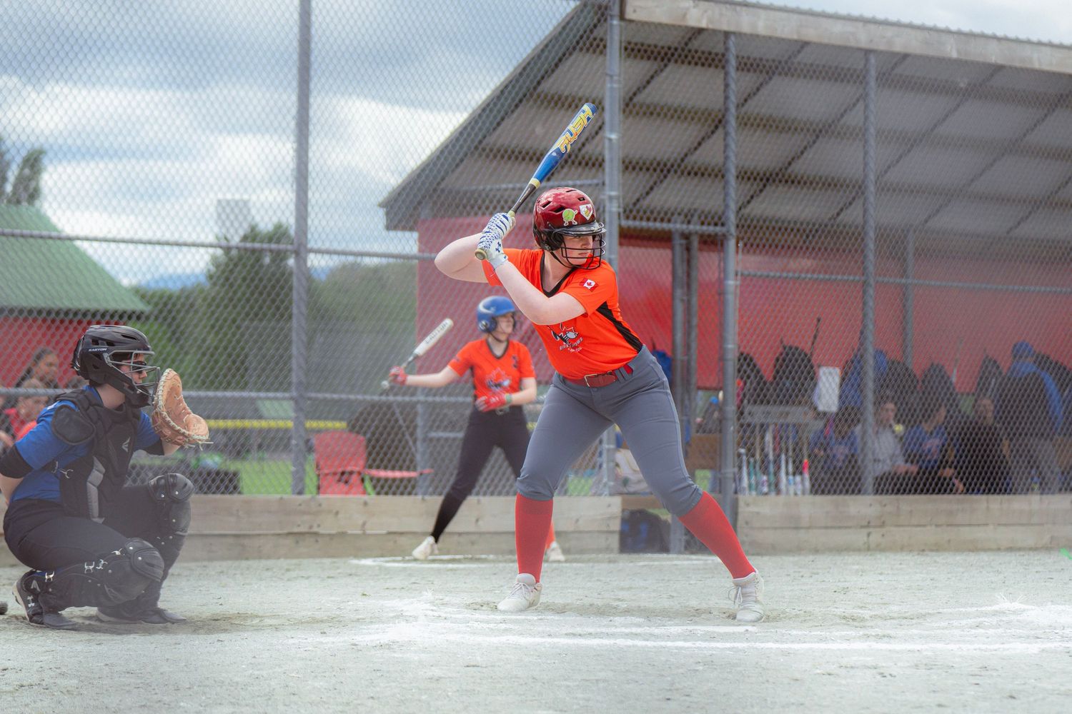 Female softball batter in orange uniform at plate with catcher crouching behind her.