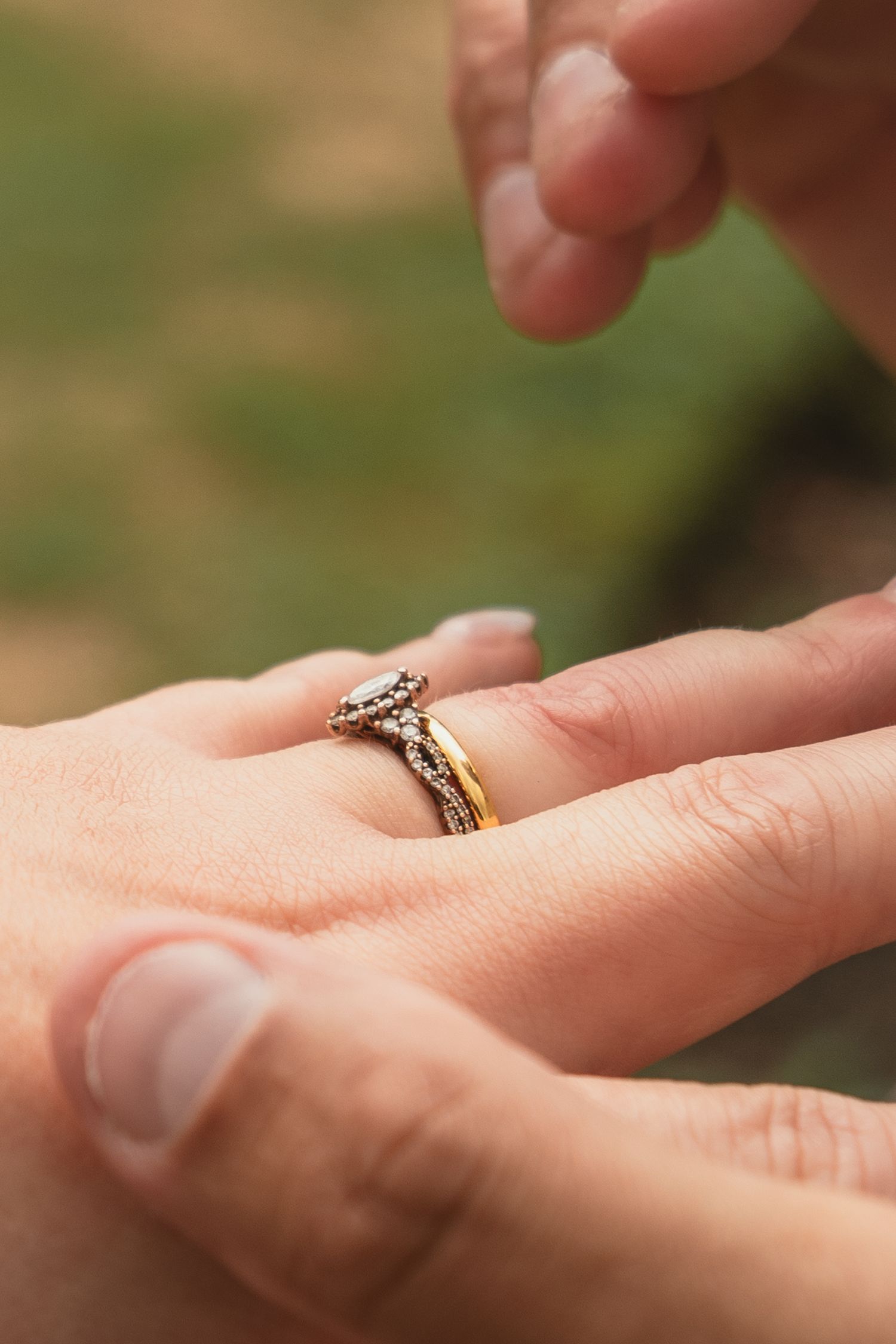 Close-up of a person placing a gold diamond engagement ring on a woman's finger outdoors.