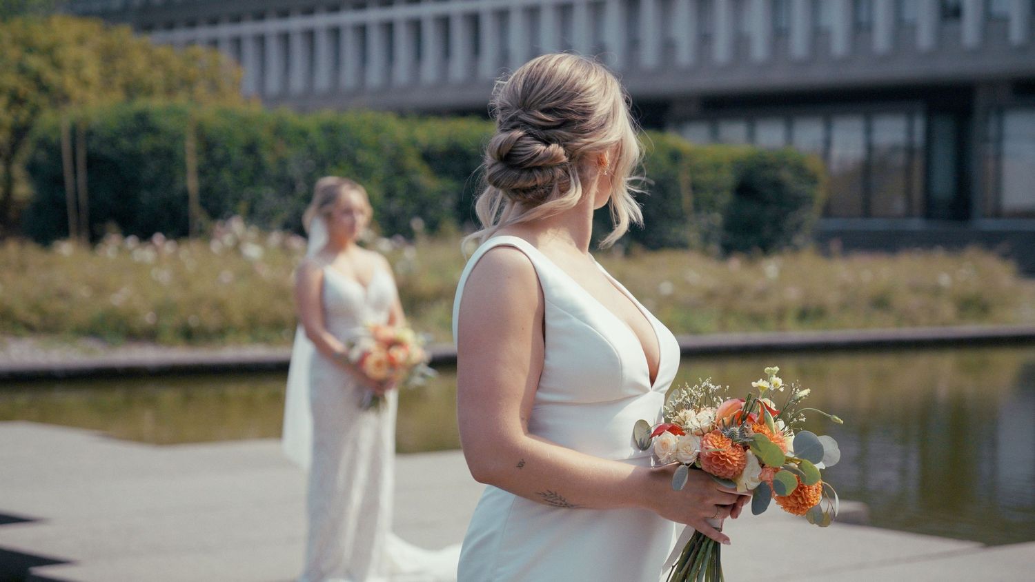 Two brides in white dresses holding bouquets of orange and white flowers pose near a fountain outdoors.