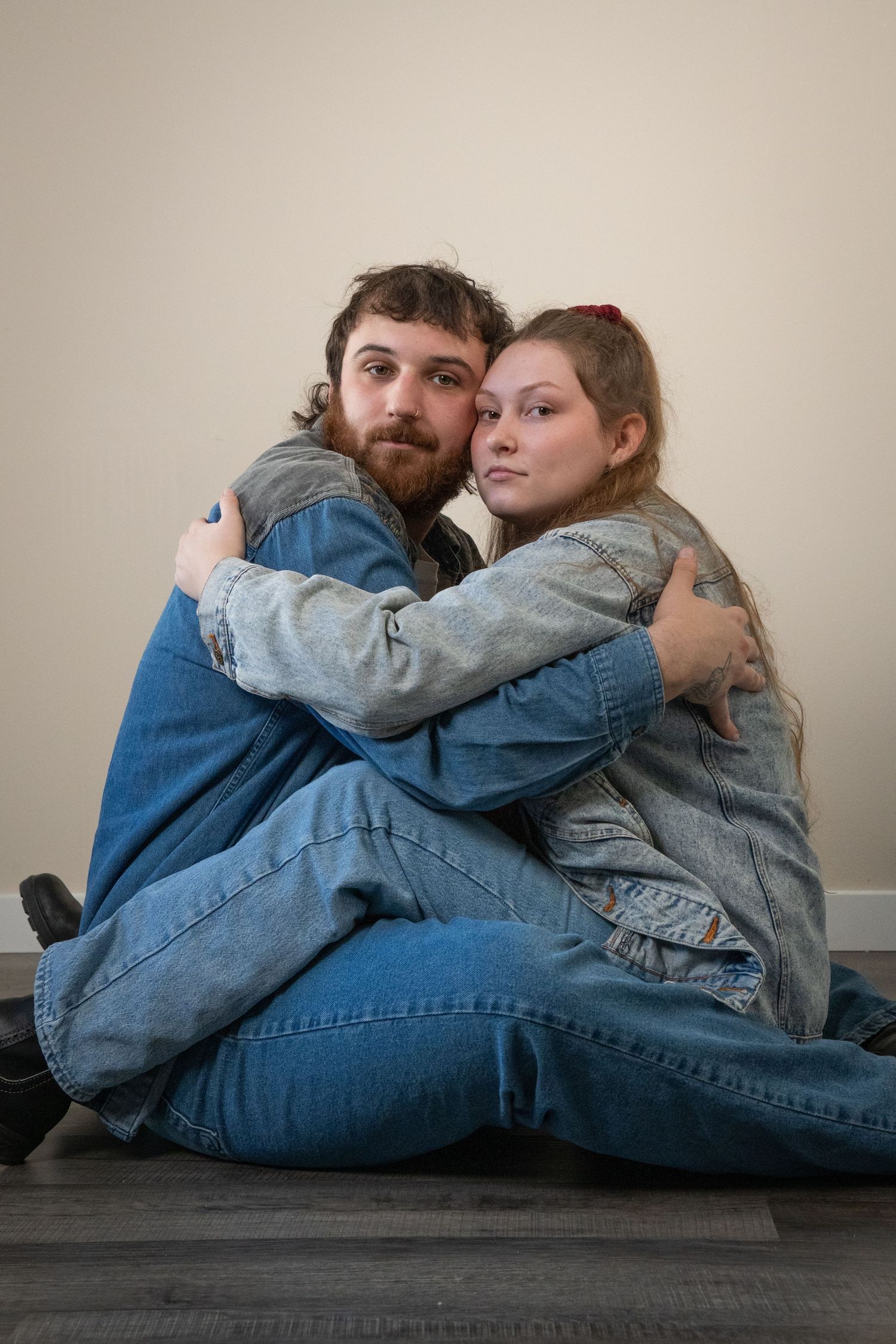 A bearded man and young woman in denim jackets share a warm embrace while sitting on a hardwood floor.