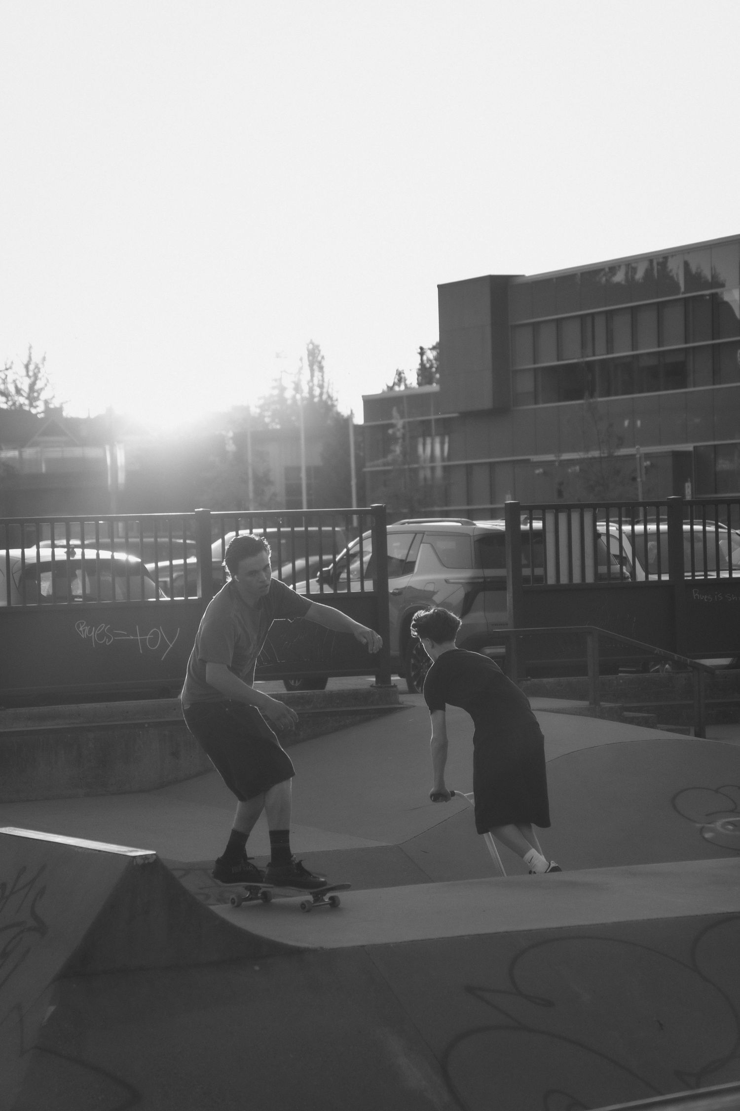 Black and white photo of two skateboarders at a skate park during golden hour sunset.