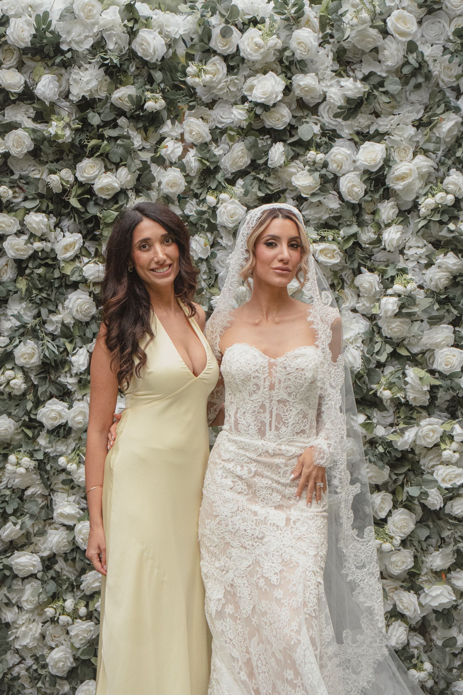 Bride in lace gown and veil poses with bridesmaid in yellow dress before white floral wall backdrop.
