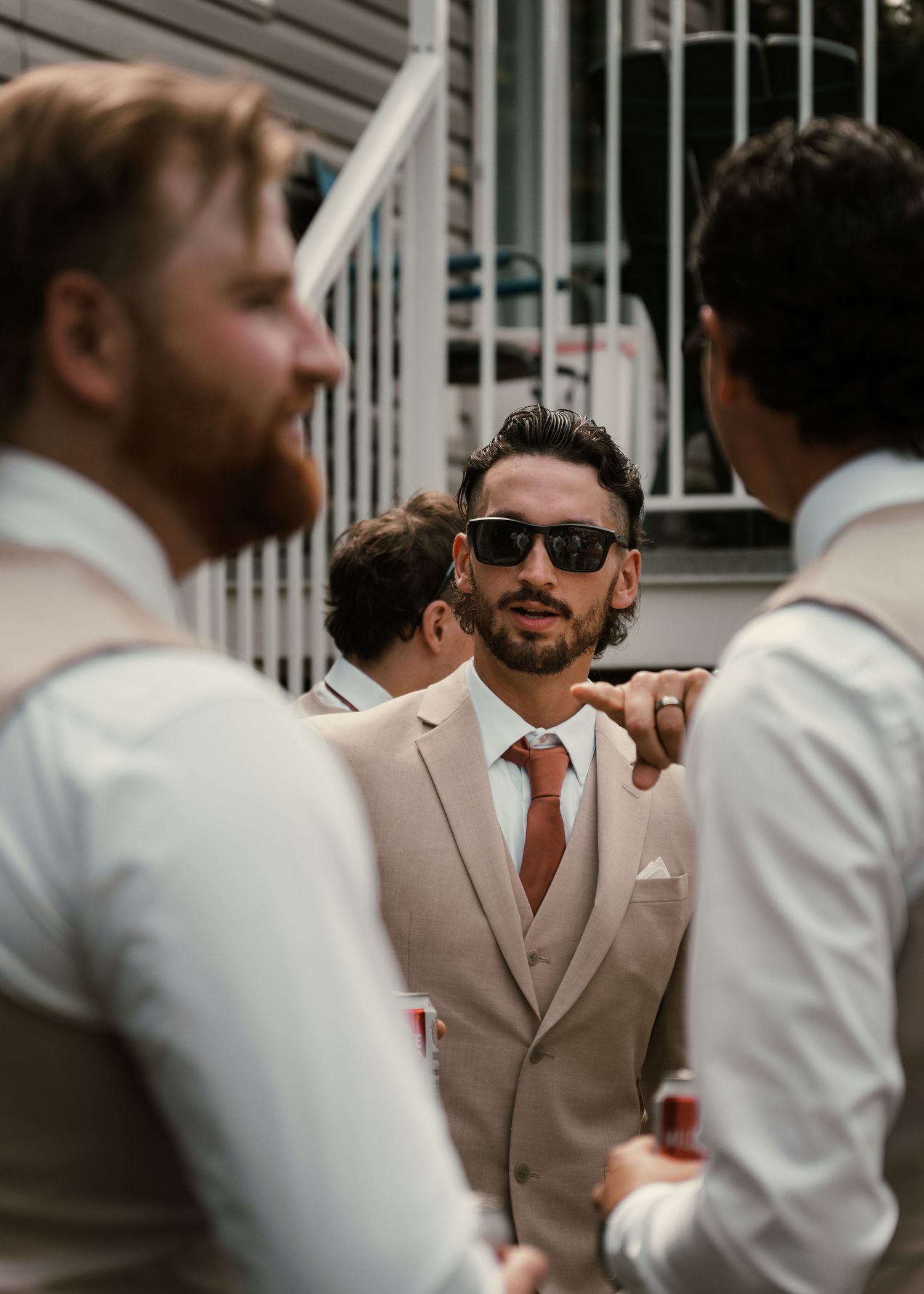 Groomsmen in tan suits and rust ties socializing outdoors before a wedding ceremony.
