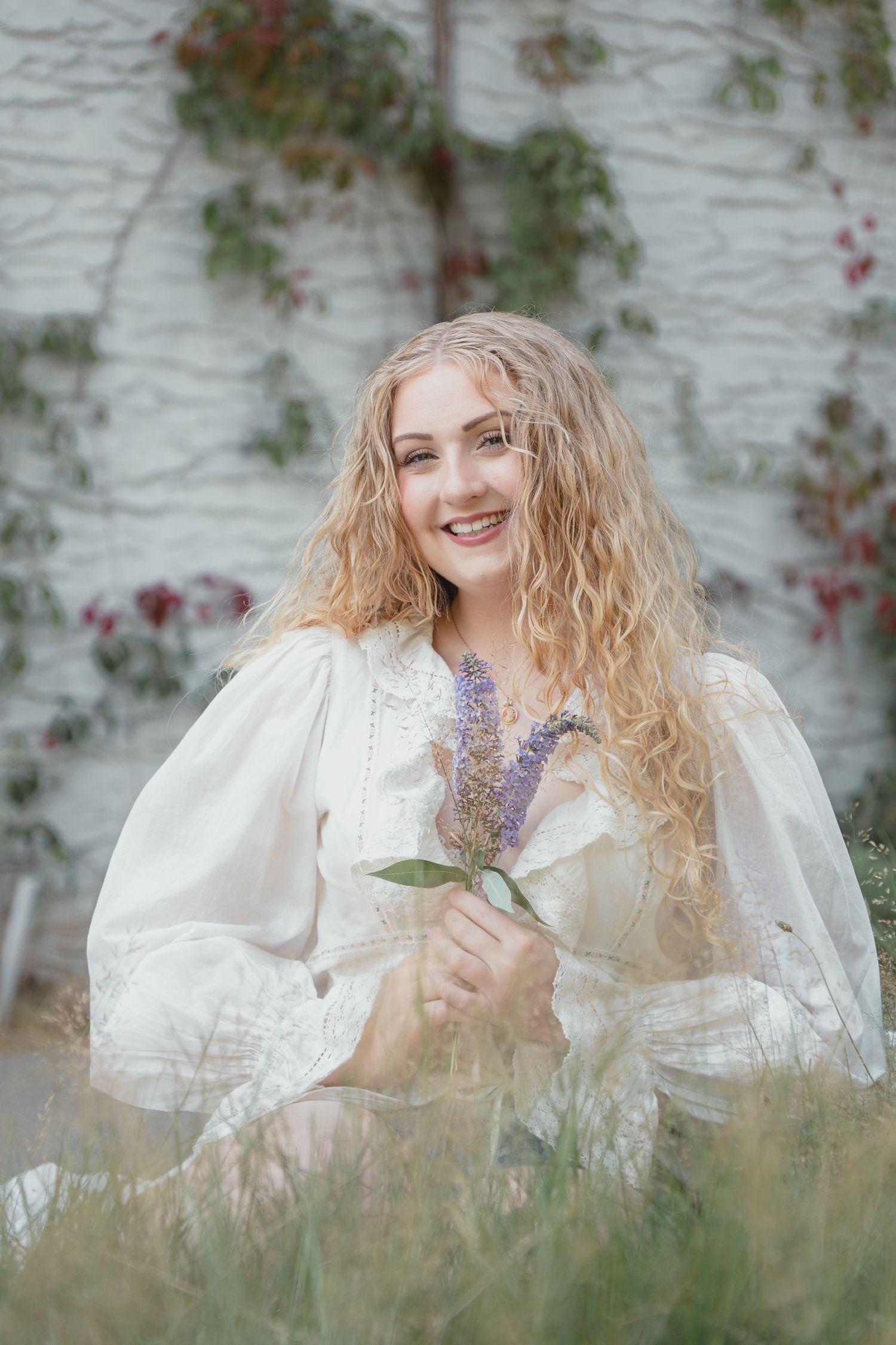 Curly blonde woman in white prairie dress sitting in tall grass holding purple lavender flowers.