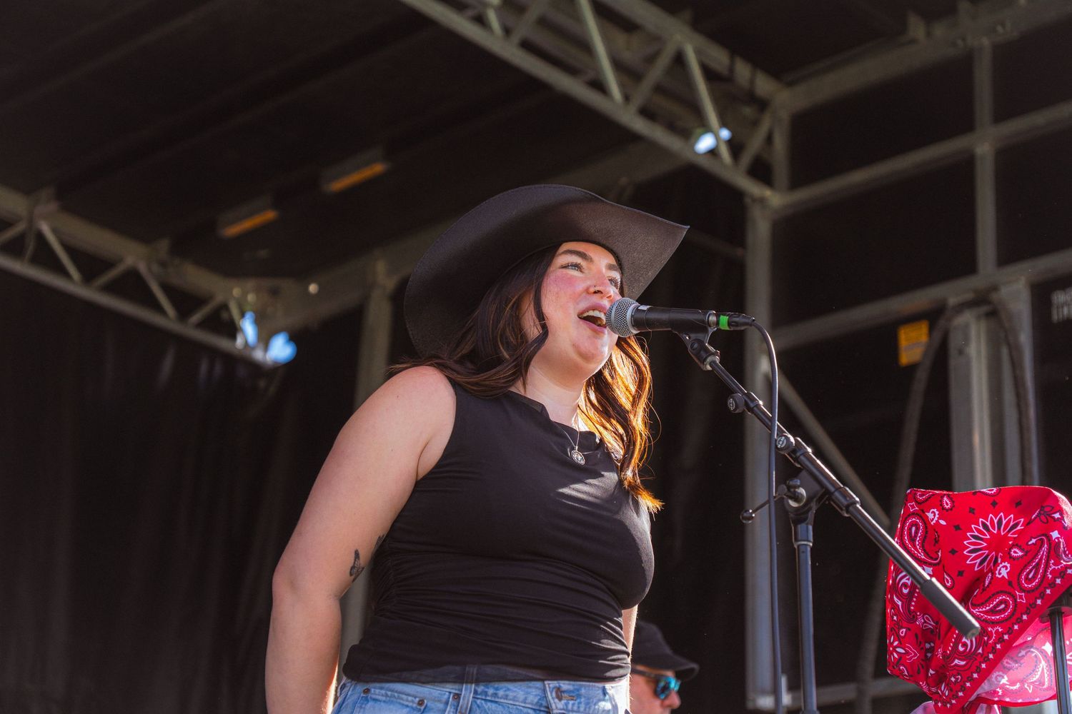 Young woman singing on outdoor stage wearing black cowboy hat and tank top at country music festival.