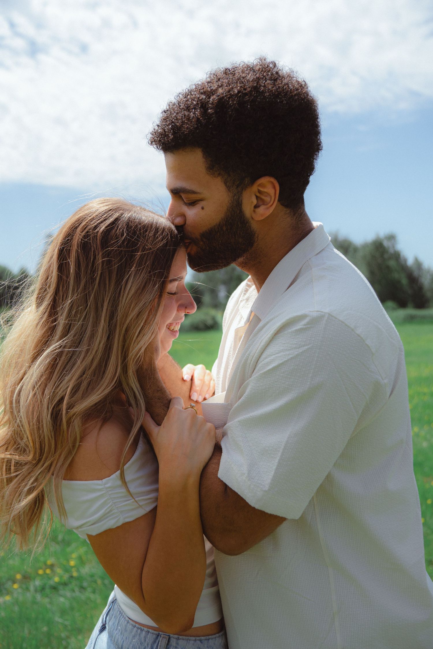 A romantic interracial couple holding hands and sharing a tender kiss on the forehead in a sunny green field.