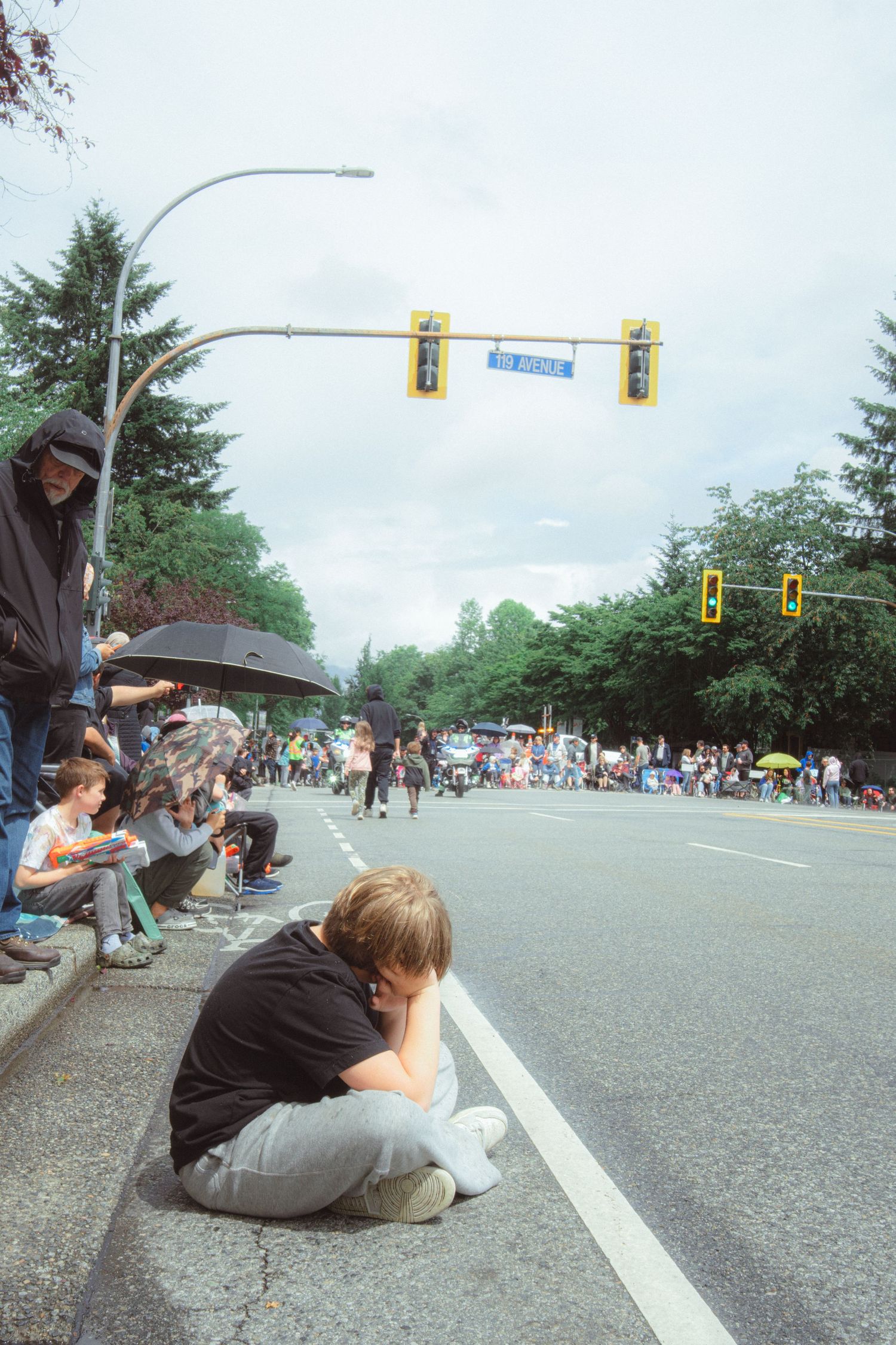Spectators line a wet street curb watching a parade on a cloudy day, with traffic lights visible overhead.