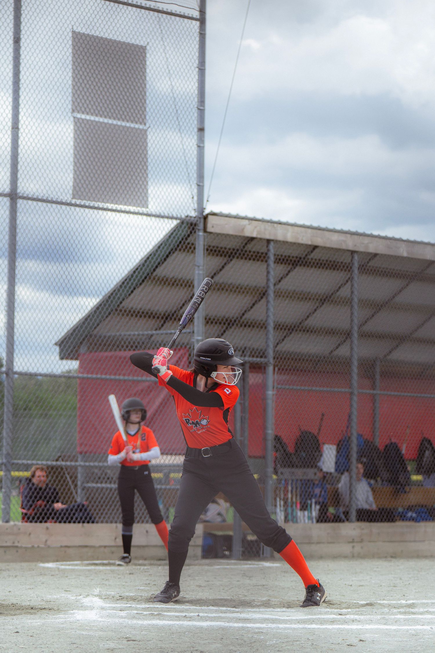 Female softball player in black and orange uniform in batting stance at home plate.