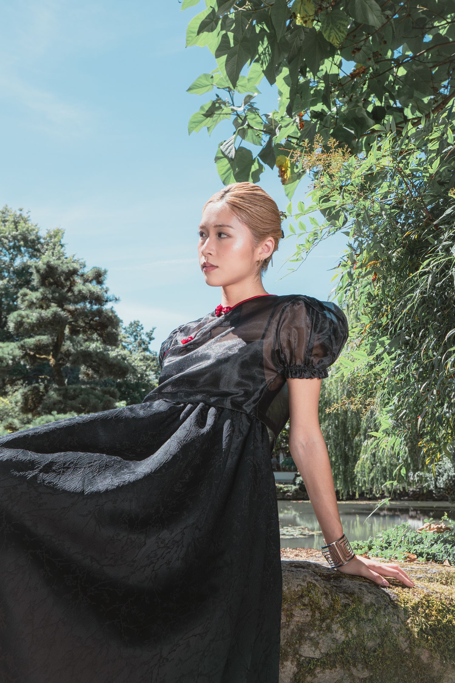 Woman in elegant black dress with red floral detail poses gracefully in a lush garden setting on a sunny day.