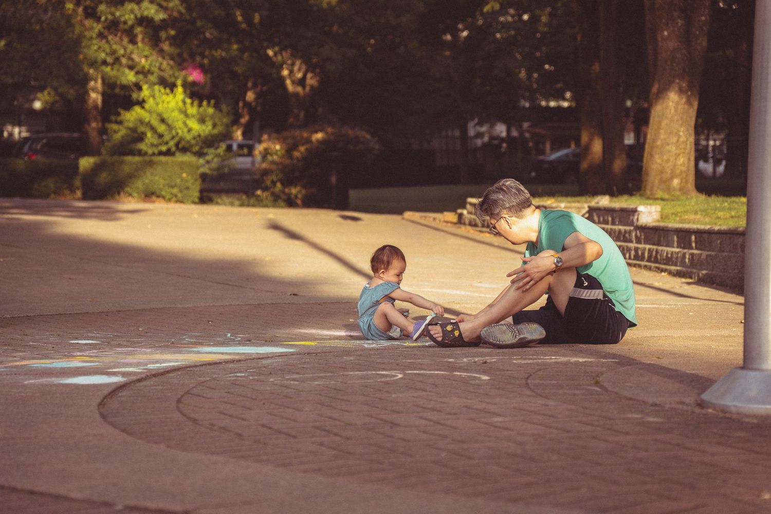 Adult and child drawing with chalk on a park sidewalk on a sunny day.